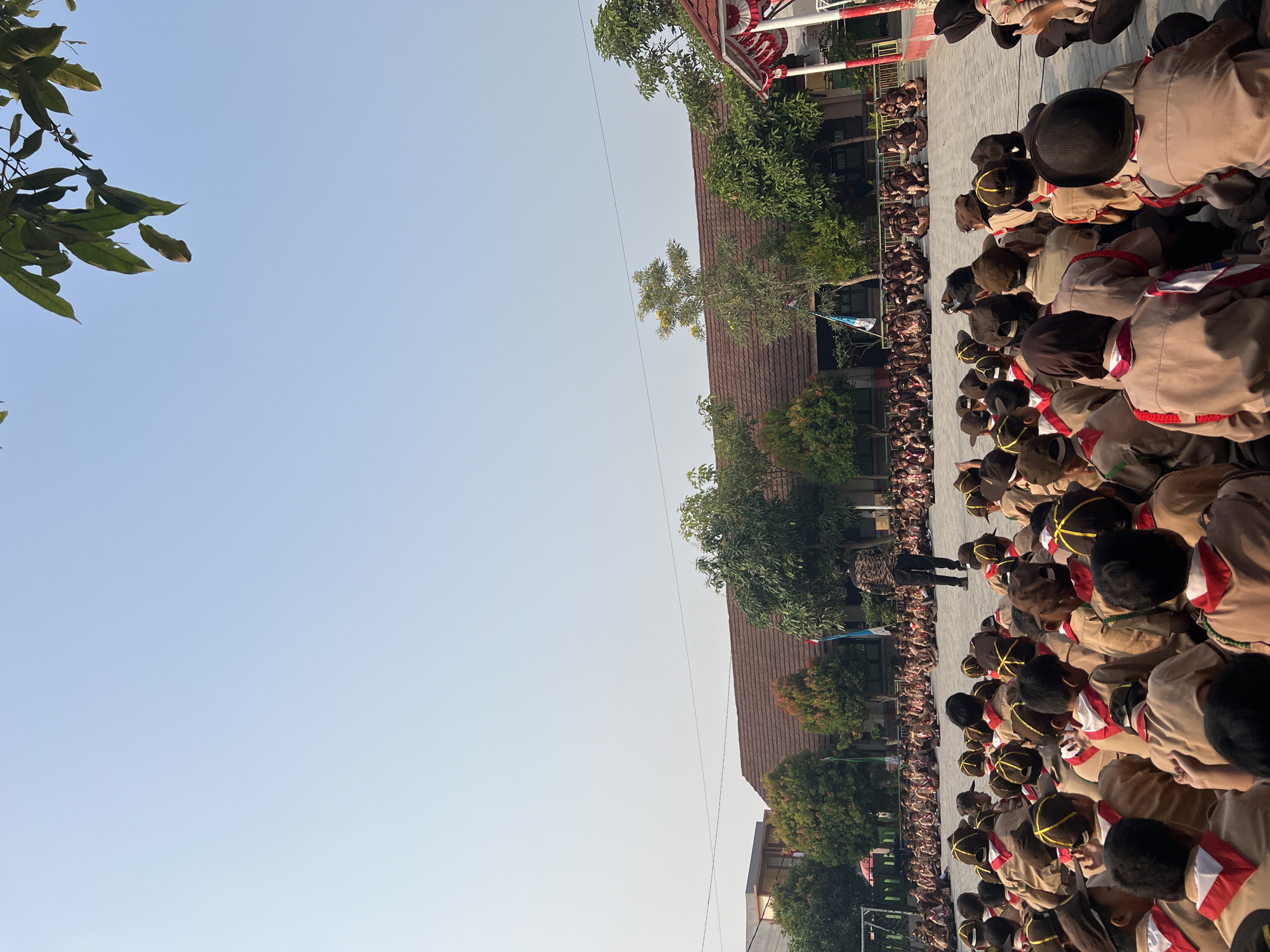 This is a medium shot, taken outdoors from a low angle, capturing a large group of children in scout uniforms sitting on the ground in what appears to be a schoolyard. The children are facing away from the camera, towards a figure standing in the distance. They are all wearing brown scout uniforms with red and white neckerchiefs and caps. Some of the children have brown caps with yellow trim, while others have plain brown caps. The ground is paved with light-colored tiles. In the background, there is a low-rise building with a brown tiled roof and trees. A red and white tent or canopy is visible on the right side of the frame. The sky is clear and blue. The location is identified as Gelam Jaya, Indonesia.