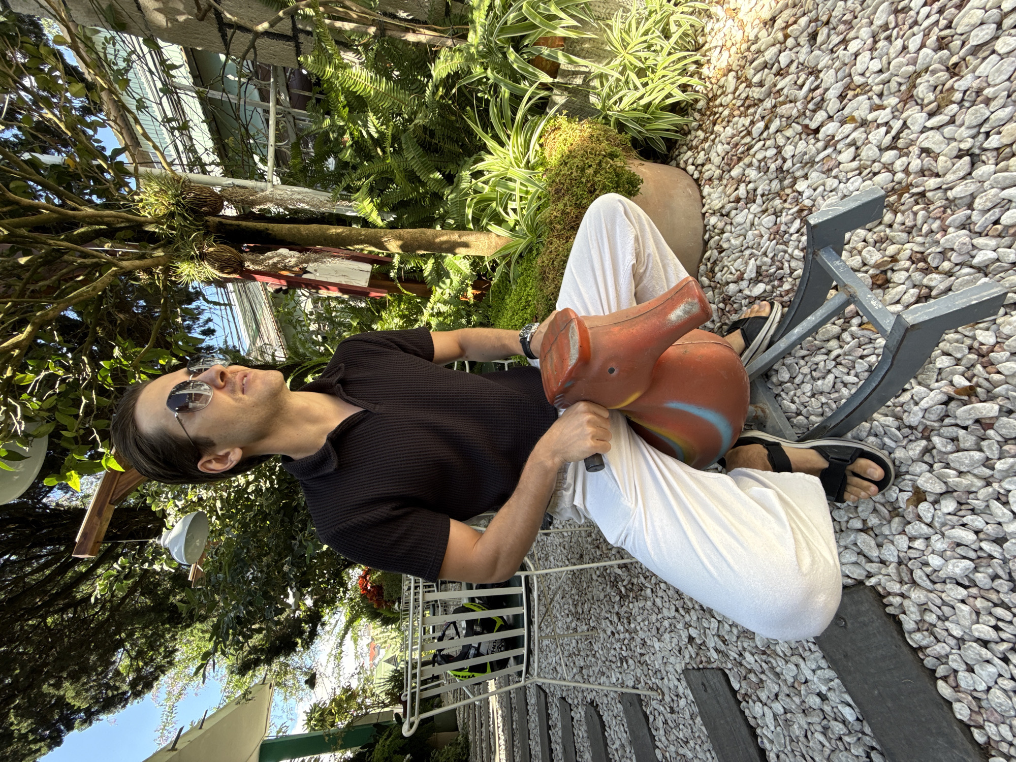 A man is sitting on a rocking horse in an outdoor area in Da Lat, Vietnam. The man is wearing sunglasses, a black collared shirt, and white pants, and black sandals. He is holding onto the handles of the rocking horse. The rocking horse is made of red and blue plastic. The area is filled with white gravel and surrounded by lush green plants. There are metal benches and stairs in the background. Overhead, there are trees and a partially obscured building.
