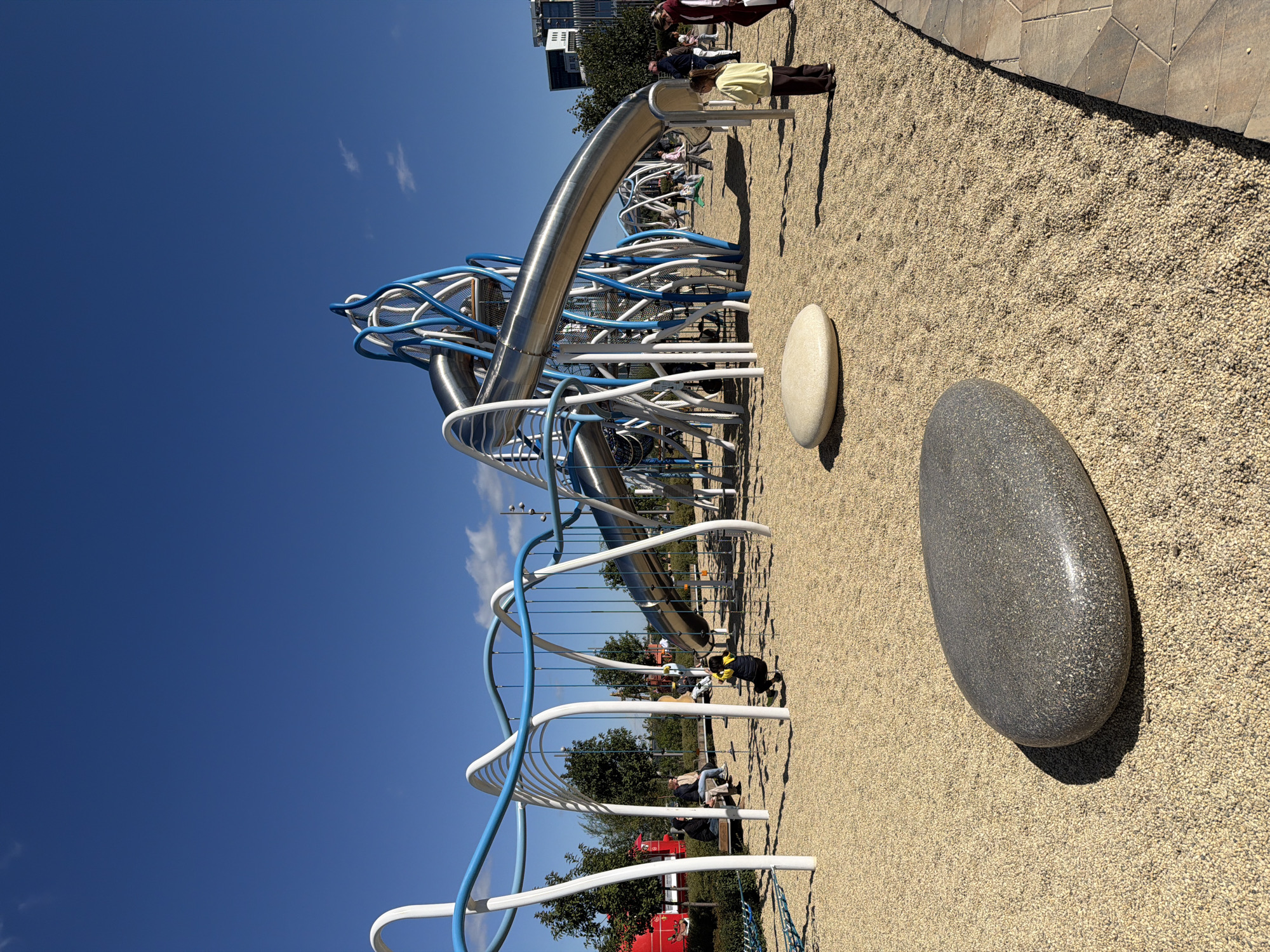 This image depicts a modern, futuristic playground in Kazan, Russia, under a clear blue sky. The playground features an elaborate climbing structure with large, spiraling metal slides and a network of blue and white tubular elements. The ground is covered with light brown, pebble-like material. Several people are visible in the image: a child is swinging on a rope swing, another child in a yellow top and brown pants stands facing away from the camera, and a group of adults are sitting on a bench in the background. A large, grey, oval-shaped boulder is prominently placed in the foreground, with a smaller, lighter-colored oval boulder behind it. In the background, trees, buildings, and a red vehicle are discernible. The overall scene suggests a vibrant and engaging recreational space.