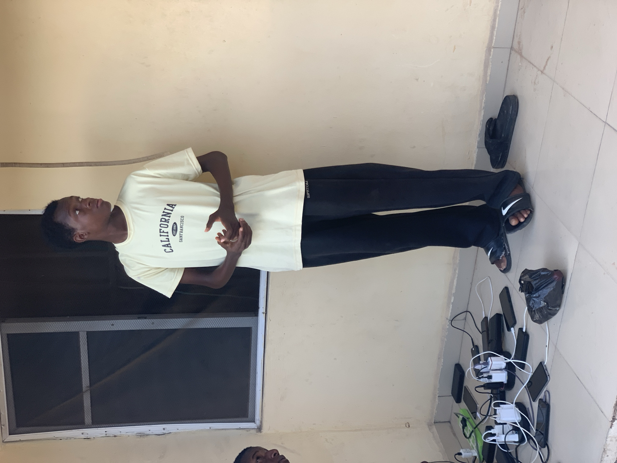 The image, taken in Lokoja, Nigeria, depicts a young Black man standing on a tiled outdoor surface, likely a veranda or porch, during daylight. He wears a light cream-colored t-shirt with the text "CALIFORNIA EST. 1997 SAN FRANCISCO" printed on it, and black trousers with "SUPERTOM" written vertically on the right leg. He is also wearing black slides featuring a white Nike swoosh. His hands are clasped in front of him, and he looks upward and slightly to his left with a neutral, contemplative expression.
To the man's right, on the tiled floor, numerous smartphones and possibly power banks are being charged. There are at least seven to eight devices connected to several white charging cables and multi-port adapters, indicating a communal charging arrangement. A black plastic bag rests near these devices, and a pair of black flip-flops is placed against the wall nearby. The background features a plain light-colored, possibly beige, wall and a window with a dark mesh screen. Part of another person's head is visible at the very bottom of the frame. The bright illumination suggests it is daytime.