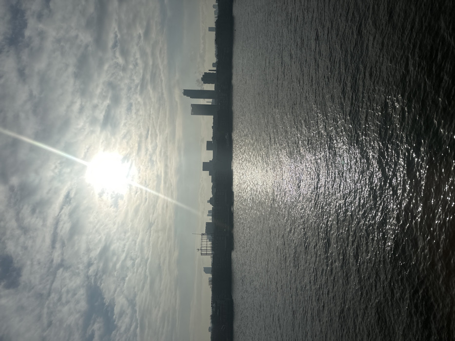 The image displays a panoramic view across the River Thames in London, United Kingdom, towards the silhouetted skyline of Canary Wharf. The scene is dominated by a bright sun in the upper-left quadrant of the frame, emitting a strong lens flare and casting a brilliant, shimmering reflection path across the ruffled dark grey surface of the river towards the lower-right. The sky above is partially covered by scattered, light-colored clouds, suggesting a moderate cloud cover.
The distant city skyline, positioned across the midground, is rendered in dark silhouette due to the intense backlighting from the sun. Prominent within this silhouette are several high-rise structures characteristic of the Canary Wharf financial district, particularly two tall towers towards the right side of the skyline cluster. To the left of these towers, a large, skeletal industrial-type framework or construction site is visible. No individuals are present in the observable scene. The overall lighting suggests either sunrise or sunset.
