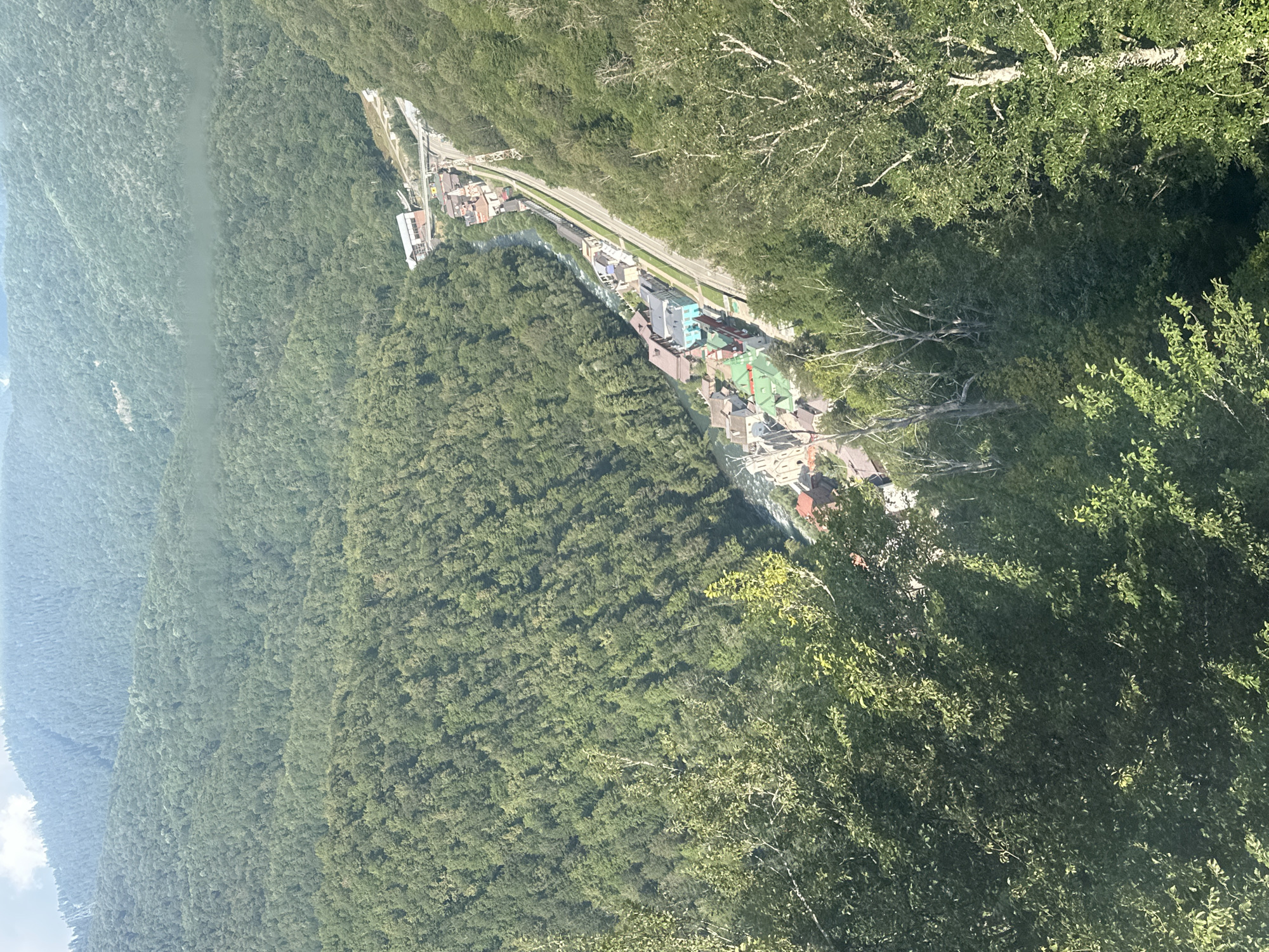This is an aerial, high-angle shot of a village nestled in a valley, with dense green forests covering the surrounding mountains. A river flows through the center of the village, with a road running parallel to it. Buildings with colorful roofs are clustered along the river and road. In the distance, the mountains fade into a hazy blue, suggesting a vast, undeveloped landscape. The foreground is filled with the leafy branches of trees, framing the scene. The location is the village of Estosadok, Russia, a mountain resort known for its ski slopes and natural beauty. The scene captures the integration of human settlement within a dramatic natural environment.