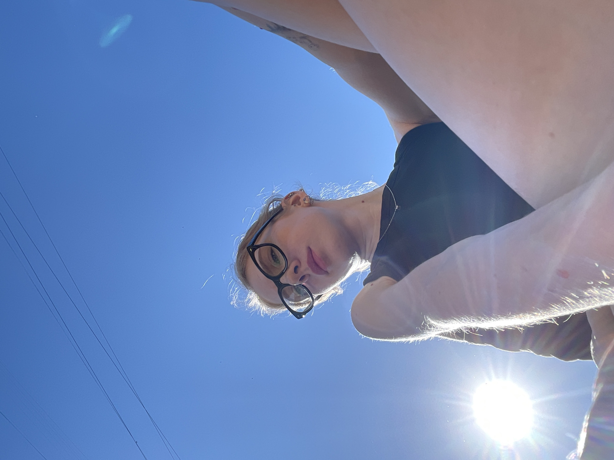 A low-angle shot of a fair-skinned person wearing black-rimmed glasses and a black t-shirt. Above the person is a clear blue sky. A bright sun is visible in the lower right of the frame, creating lens flare. There are power lines visible in the lower left. The location in Izhevsk, Russia is not discernible from the image.