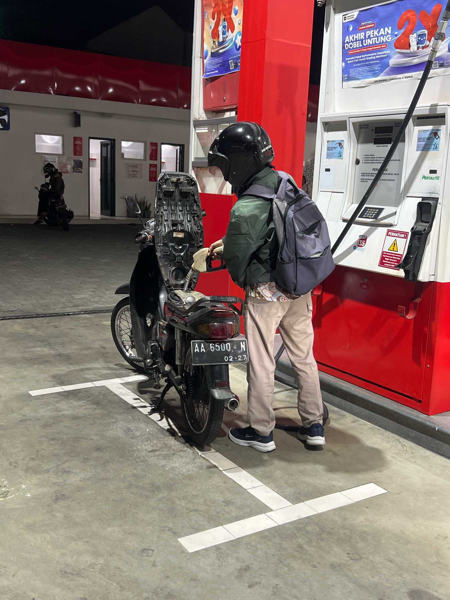 A person wearing a black helmet, green jacket, and tan pants is standing at a Pertamina gas station in Magelang, Indonesia. The person is holding a fuel nozzle and refueling a black motorcycle. The motorcycle has a license plate that reads "AA 6500 N 02-27". In the background, another person is sitting on a motorcycle. The gas station appears to be open, with lights illuminating the area. The ground is concrete, and there are white lines marking parking spaces.