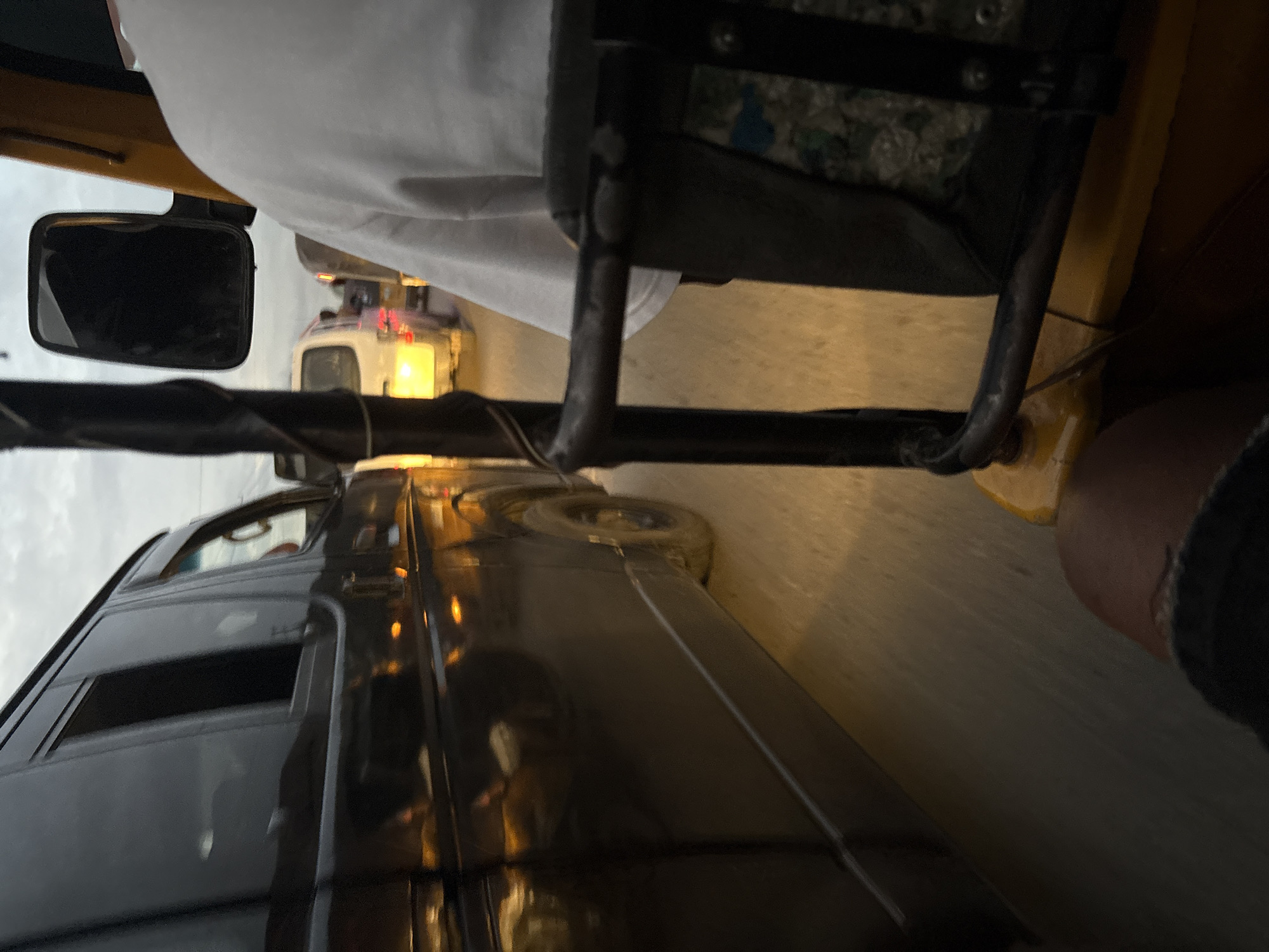 The image depicts a view from inside a yellow auto-rickshaw traveling on a street in Onitsha, Nigeria. The auto-rickshaw is in motion, indicated by the blurred road and the reflections on the dark van in the foreground. The van, a black commercial vehicle, is in the adjacent lane, also appearing to be in traffic. Its windows are dark and reflect the ambient light. Further ahead, a white commercial van is visible, its headlights illuminated. The sky is overcast, suggesting it might be late afternoon or evening. The overall scene is one of moderate traffic in an urban environment.