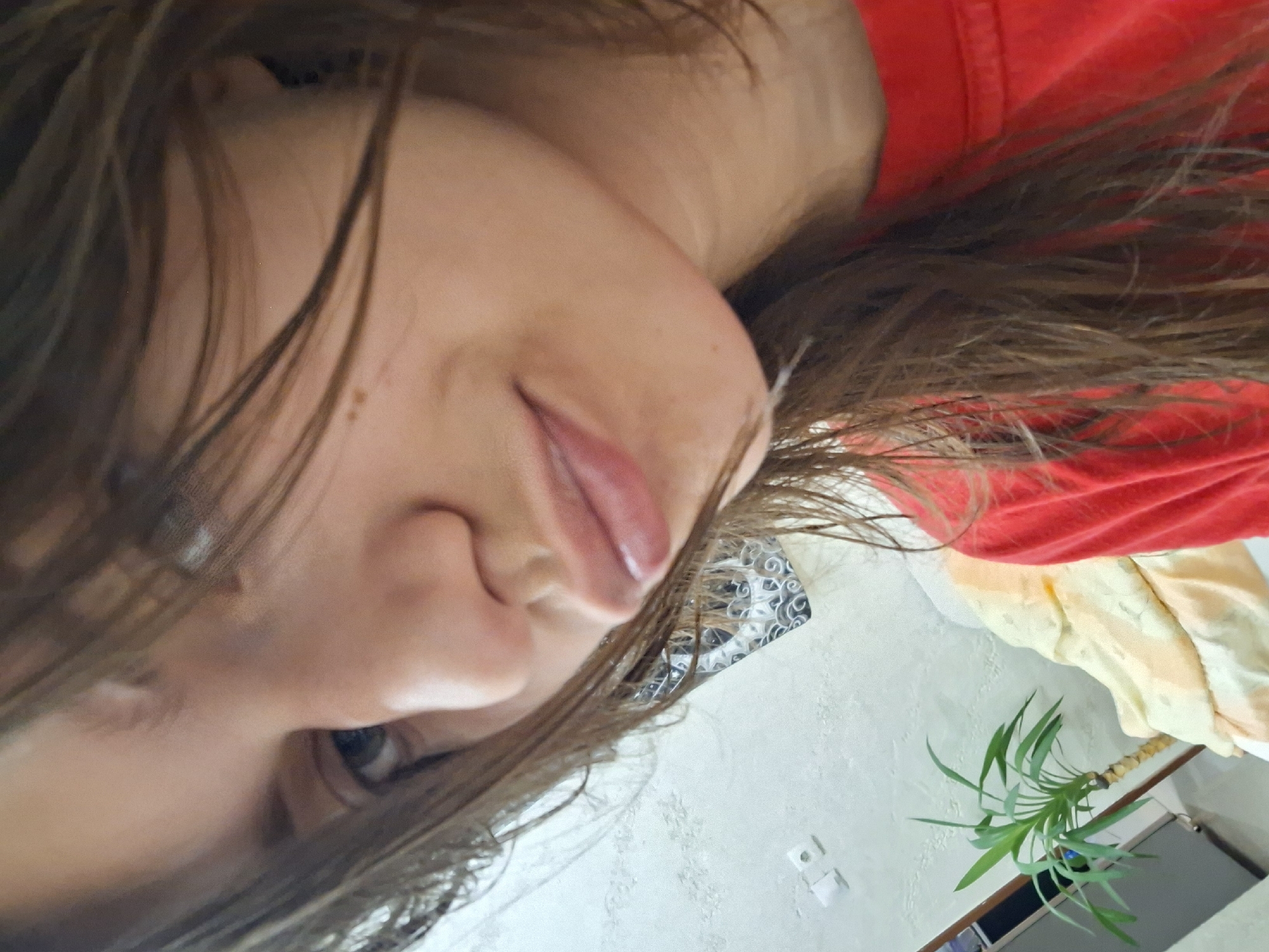 A person is indoors, viewed at an angle. The person has light skin, dark hair, and is wearing a red shirt. The background includes a white wall with a decorative pattern, an electrical outlet, a plant in a pot, and a glimpse of what appears to be furniture. I cannot provide location in the city of Panagyurishte, Bulgaria.