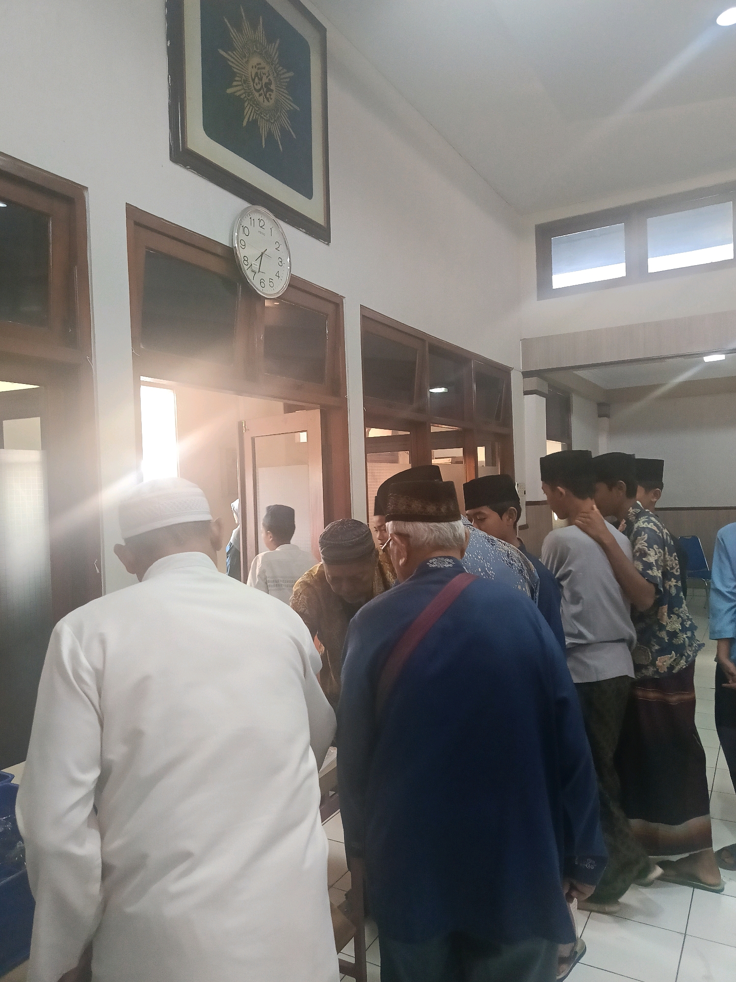 In a brightly lit indoor space, possibly a community hall or mosque in Yogyakarta, Indonesia, several men are gathered. A white analog clock is mounted on the wall, indicating a time between 3:15 and 3:20. Above the clock, a framed emblem with intricate golden detailing is displayed. The lighting creates lens flare effects coming from the right side of the frame.
In the foreground, the back of a man wearing a white shirt and a white peci (a type of cap) is visible. He appears to be looking towards an open doorway. To his right, another man in a dark blue jacket with a red strap across his shoulder, also wearing a peci, is facing away from the camera and engaging with others.
A group of men and young men are congregating in the middle ground, some wearing peci hats and patterned shirts or sarongs. The doorway leads into another room, where the bright light suggests an exit or another well-lit area. The floor is tiled with light-colored square tiles. The overall atmosphere appears to be one of a gathering or social interaction.