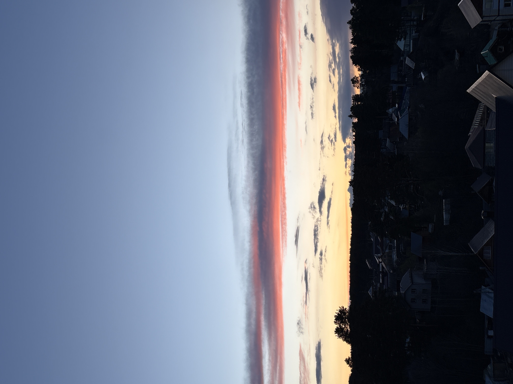 This image depicts a serene twilight landscape, likely a sunset or sunrise, over a settlement in Chaikovsky, Russia. The sky dominates the upper portion of the frame, transitioning from a clear, deep blue at the top to lighter hues and vibrant colors closer to the horizon.
A striking feature is the long, layered cloud formation stretching across the middle of the sky. This cloud band displays intense shades of dark red, orange, and pink, interspersed with lighter grey and white sections, suggesting the sun's low angle. Below this, the sky near the horizon glows with soft yellows and pale oranges, accented by scattered, darker cloud wisps.
The lower third of the image is characterized by a dark silhouette of a landscape. A dense line of trees forms the immediate horizon, behind which numerous houses and buildings are visible as dark, undefined shapes against the colorful sky. Various rooftops, some flat and others gabled, protrude as distinct geometric forms within the shadowed foreground. The overall impression is one of low light, with minimal detail discernible in the structures or vegetation, emphasizing their outlines against the vibrant sky.
No human activity or visible text is present in the image. The scene evokes a quiet, peaceful atmosphere at the close or beginning of the day in a rural or suburban setting.