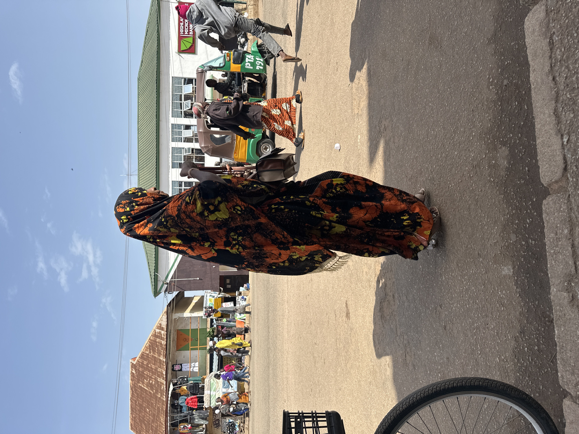 In Bukuru, Nigeria, on a paved street, a woman wearing a brightly colored headscarf and dress is walking. Behind her, several other people are walking in the same direction, with a group of vendors in stalls to the left. In the background, a green and white building with the "Highland Microfinance Bank" logo is visible, along with a three-wheeled auto-rickshaw. The scene is illuminated by sunlight, casting shadows on the street.