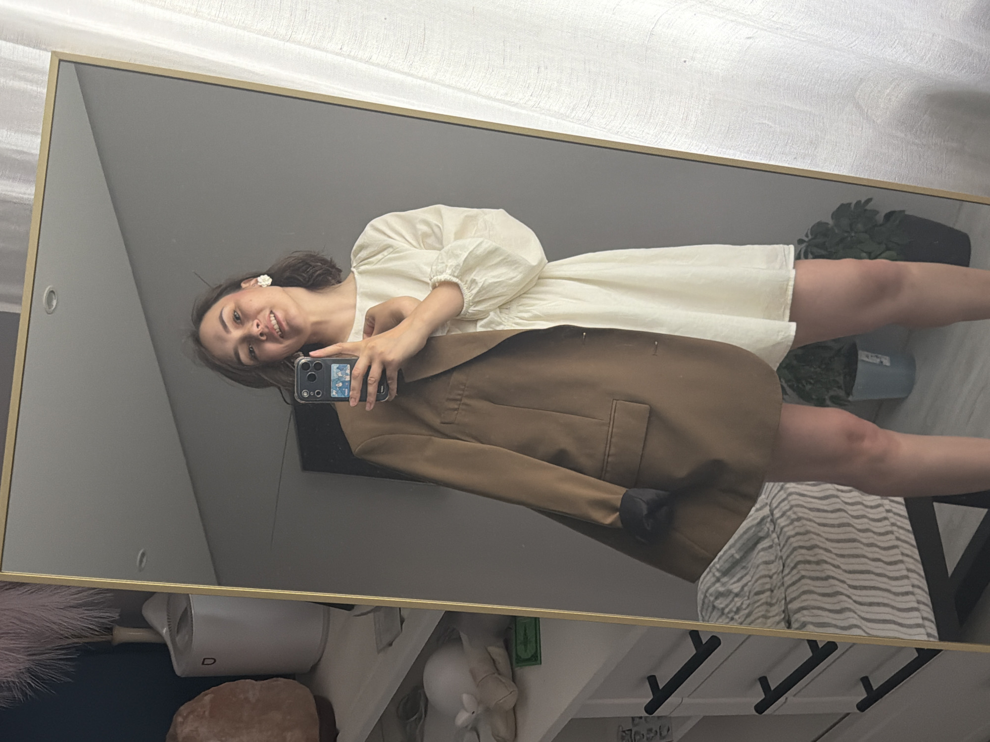 The media file is a vertical, full-body mirror selfie taken by a young woman. She is smiling and looking at the camera, holding up her smartphone to capture the reflection. She is wearing a cream-colored puff-sleeved dress and a brown blazer. The setting appears to be a room with a large, gold-framed mirror dominating the frame. In the background, reflected in the mirror, are shelves with various objects, including decorative items, plants in pots, and striped fabric. The lighting suggests it is daytime. The woman's expression is happy and relaxed.