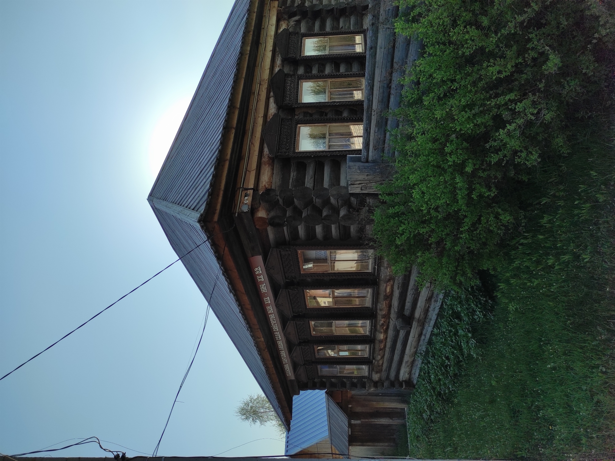The image depicts a wooden house in Chaikovsky, Russia. The house has a metal roof and decorative window frames. There are green bushes growing around the side of the house. There are power lines extending from the bottom left of the frame to the roof of the building. The sky is light blue and bright. The sign on the house is written in Cyrillic.