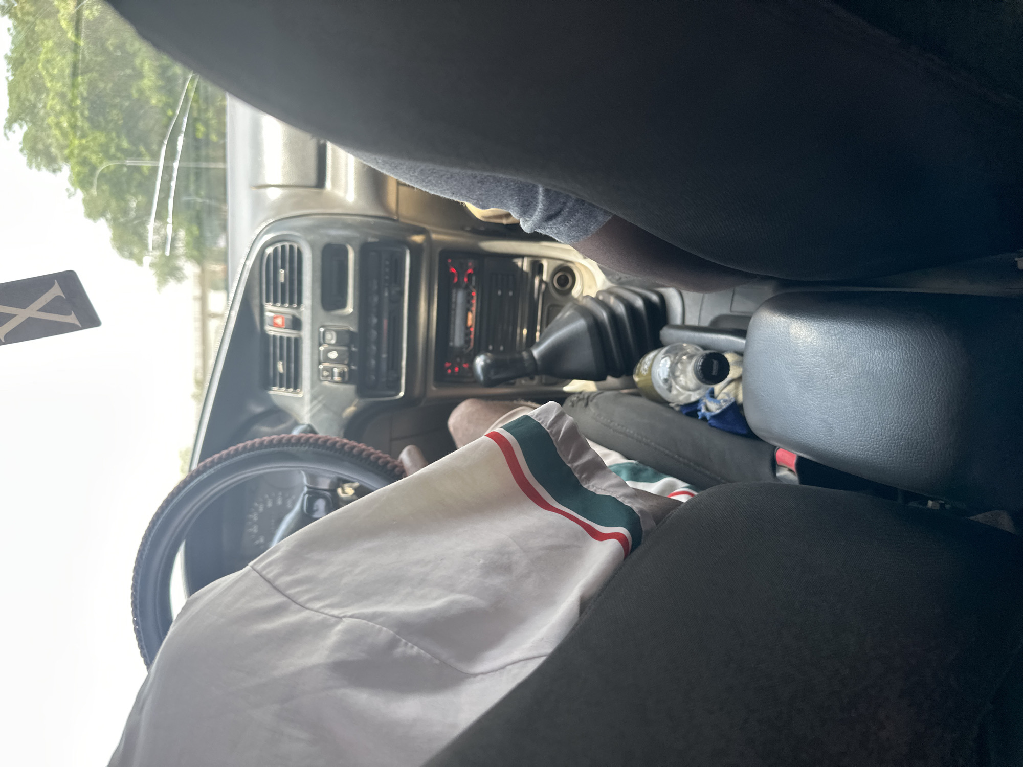 The image shows the interior of a vehicle, likely a truck or SUV, from the passenger's perspective looking towards the dashboard and driver's seat. The orientation of the image is vertical, with the top of the frame appearing to be the sky outside the windshield.
In the foreground, the driver's seat and part of the steering wheel are visible. A white garment with red and green stripes is draped over the armrest or seat. The dashboard features a radio with glowing red lights, air vents, and other controls. A gear shift lever is prominent, with a black rubber boot. A clear plastic bottle of water is also visible on the center console.
Outside the windshield, trees and a street light are visible through the glare, suggesting it is daytime, possibly morning or late afternoon. The lighting suggests a bright day. The overall scene appears to be a moment of travel within Abuja, Nigeria. There is no discernible emotion conveyed by the composition.
A black sticker with a white "X" symbol is attached to the windshield on the left side.