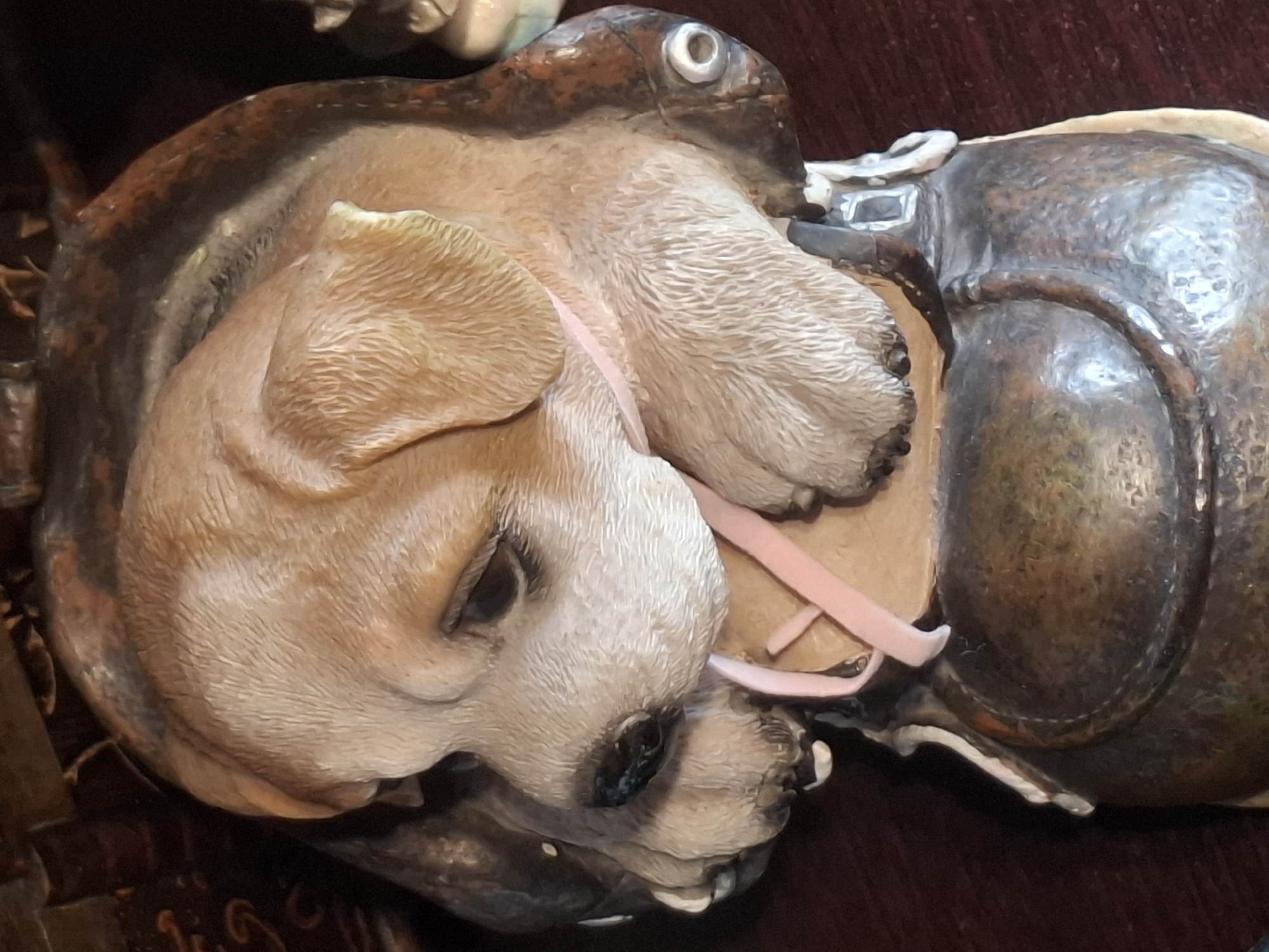 The image shows a close-up, dimly lit, overhead view of a decorative statue of a puppy wearing a collar. The puppy is light brown and beige with textured fur, and it appears to be looking down and to the left. A thin, pale pink ribbon is tied around its neck and drapes over its paws. The puppy is part of a larger, dark brown, metallic-looking object that resembles a helmet or a piece of armor. The statue is placed on a dark, wooden surface, likely a table or shelf. The background is dark and indistinct, with hints of other objects, possibly books or other decorative items, suggesting an indoor setting. There are no people or actions depicted in the image. The image is not located in the city of Almaty, Kazakhstan.