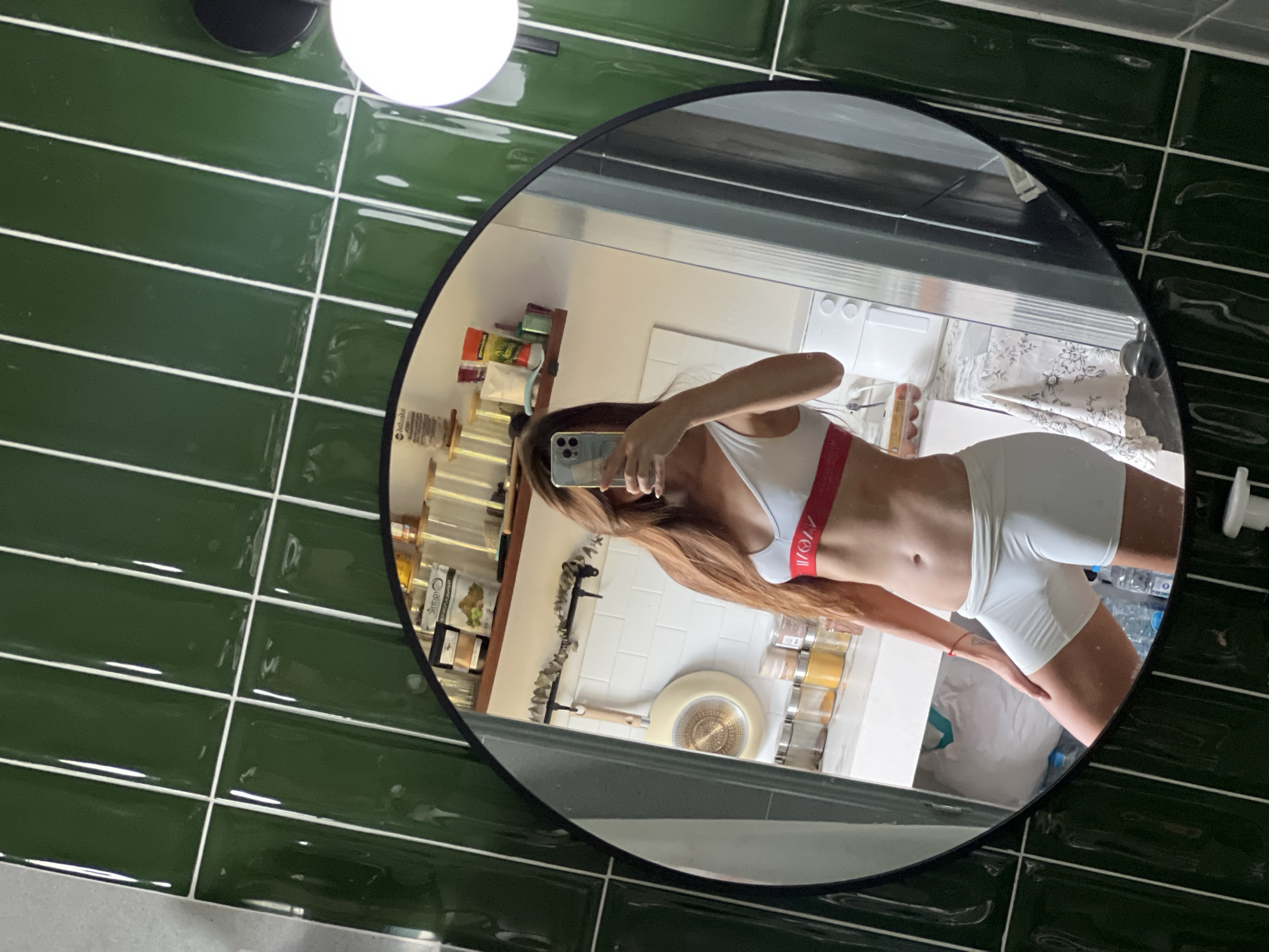 A mirror reflects a woman in athletic wear taking a selfie in a bathroom. The mirror is round with a black frame, positioned against a green tiled wall. The woman is wearing a white sports bra with a red stripe and white shorts. The background reflected in the mirror includes shelves with various items, a white counter, and a sink area. The location and city are unspecified.