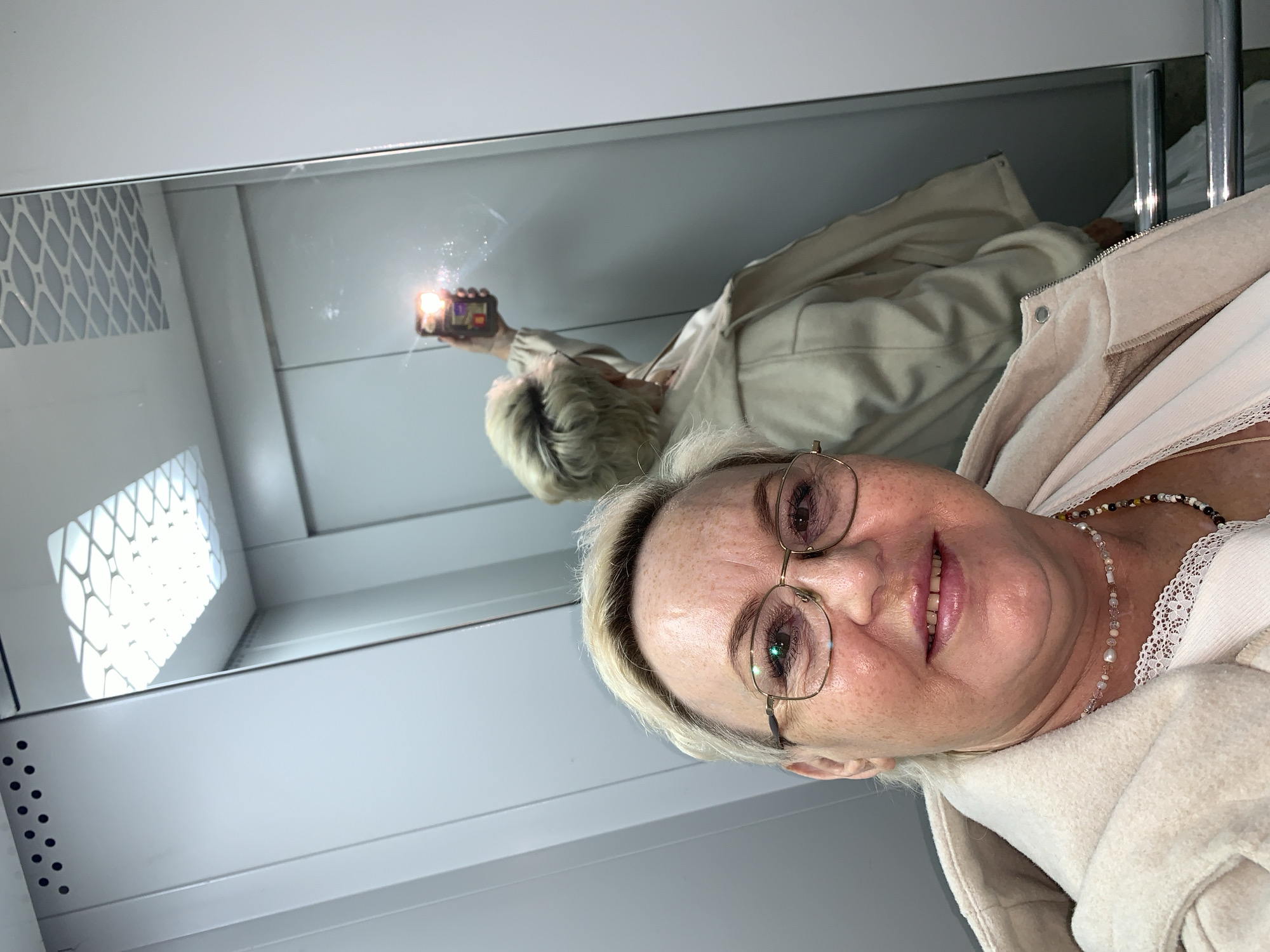 This image captures a mirror selfie of an older woman in an indoor setting, likely in Chaikovsky, Russia. The woman, with light blonde hair and wearing glasses, is visible in the foreground from the chest up, looking directly at the viewer with a gentle smile. She is dressed in a light-colored, possibly cream or beige, hooded jacket or robe worn over a top with delicate lace trim, and a multi-colored beaded necklace.
In the upper part of the frame, a large mirror reflects her entire upper body as she holds a smartphone, its flash brightly illuminated, indicating the act of taking the selfie. The reflection shows her full attire and the light-colored, possibly white or light gray, walls of the room. Distinct features visible in the mirror's reflection include a patterned grate or vent on the left side and some metal bars on the right, which could be part of a bed frame or other furniture. The overall aesthetic of the room suggests a clean, modern, and possibly institutional environment, such as a hospital room or a contemporary living space. No visible text is present in the image.