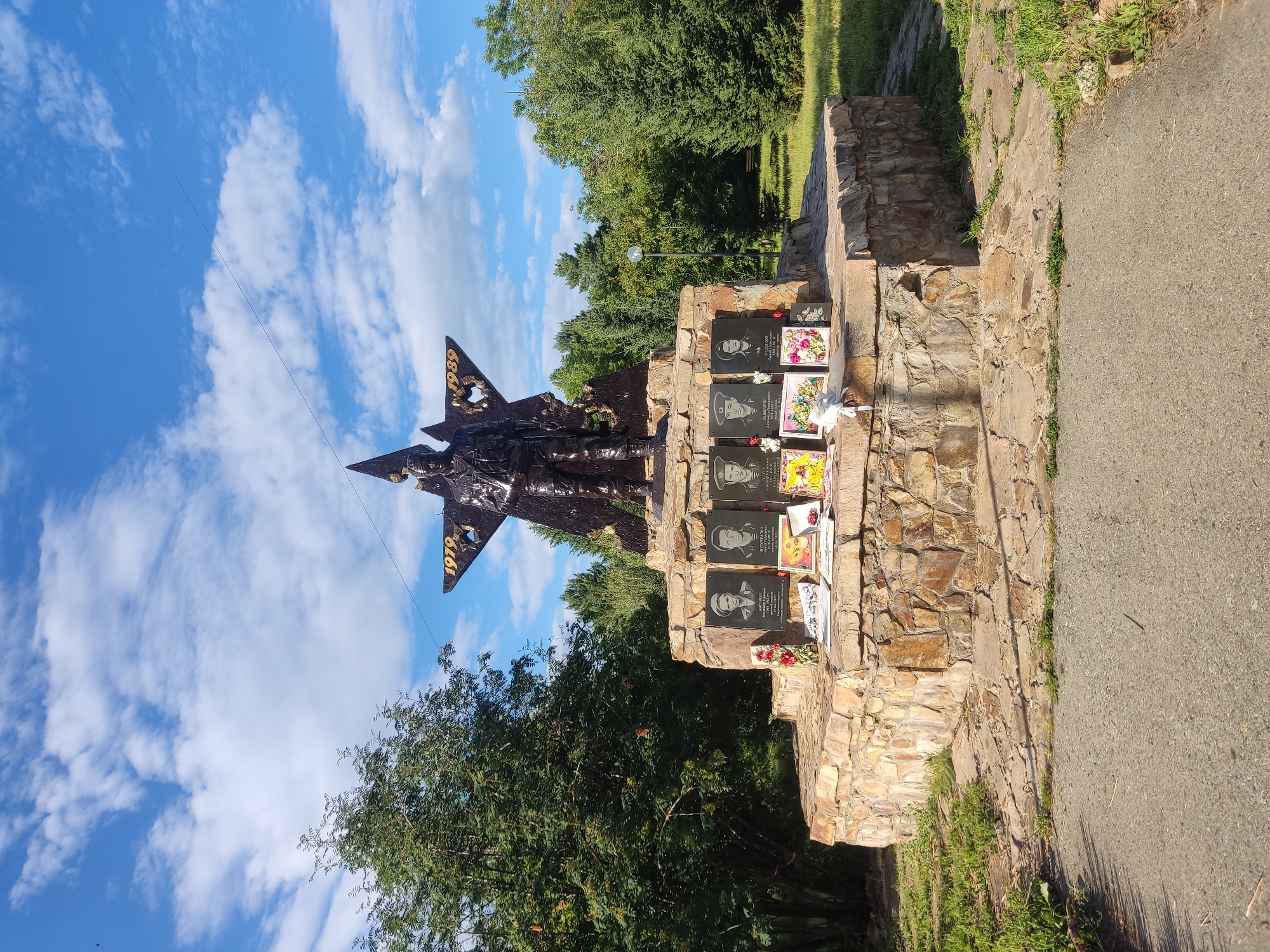 A war memorial is depicted in an outdoor setting on a sunny day. The memorial features a large, dark, star-shaped structure with a soldier figure standing atop it. The years "1979" and "1989" are visible on the star. The base of the memorial is constructed from rough-hewn stones, forming a circular platform. Mounted on this stone base are several black plaques, each displaying a portrait of a person and text below. Several floral arrangements and framed pictures are placed on the platform in front of the plaques. Surrounding the memorial is a grassy area with scattered trees and a paved walkway in the foreground. The sky above is blue with scattered white clouds.