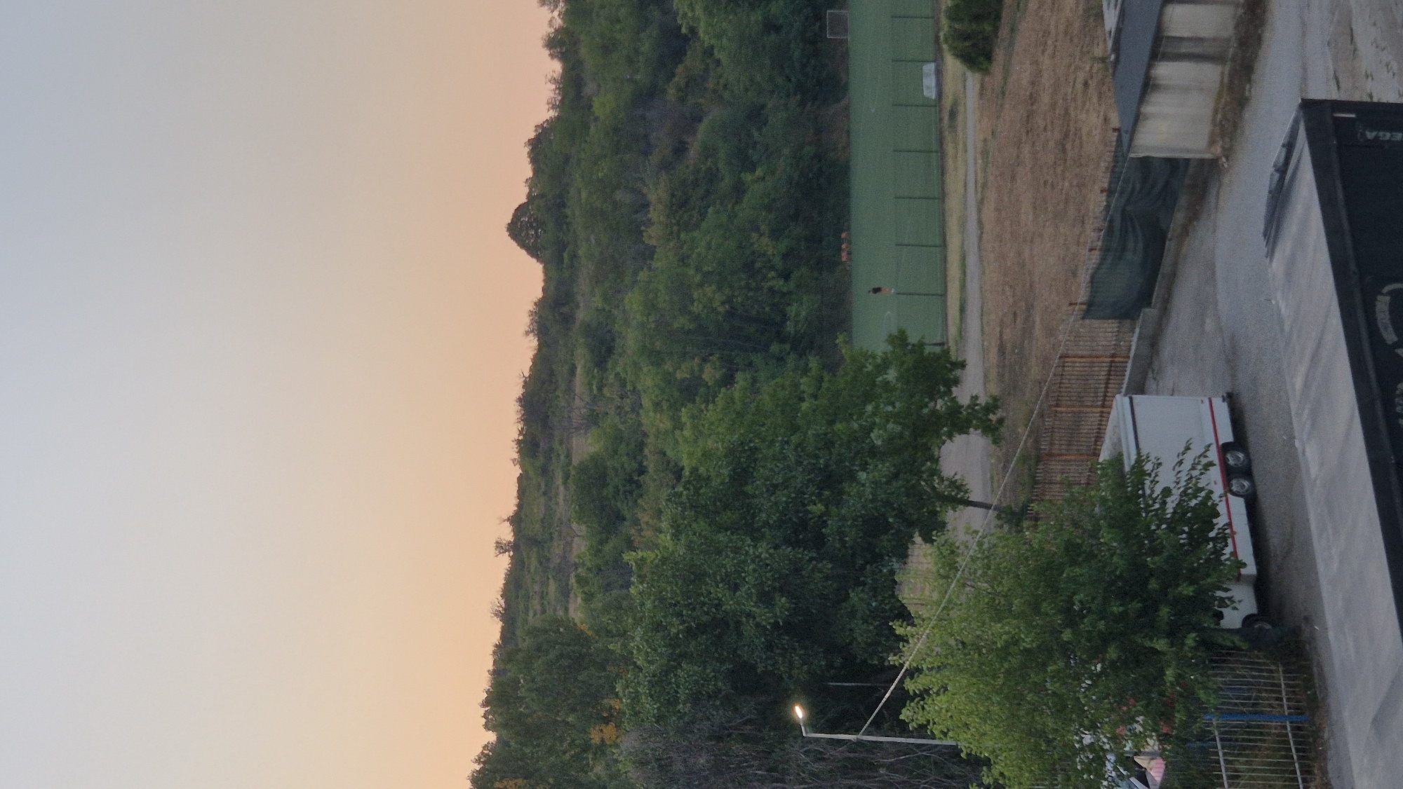 The image shows a view of a hilly, tree-covered landscape with a soccer field in the middle ground. The sky is a soft gradient of pale blue to peach, indicating either dawn or dusk. In the foreground, there is a white trailer with red trim and dual axles parked on a paved surface. To the left of the trailer, a street lamp with a bright light is visible. A dark, rectangular structure is also in the foreground, partially obscuring the view of the trailer. A green mesh fence runs along the perimeter of a section of the yard. On the soccer field, two figures are visible, one in the distance and one closer, both appearing to be playing soccer. The general environment suggests an outdoor recreational area.