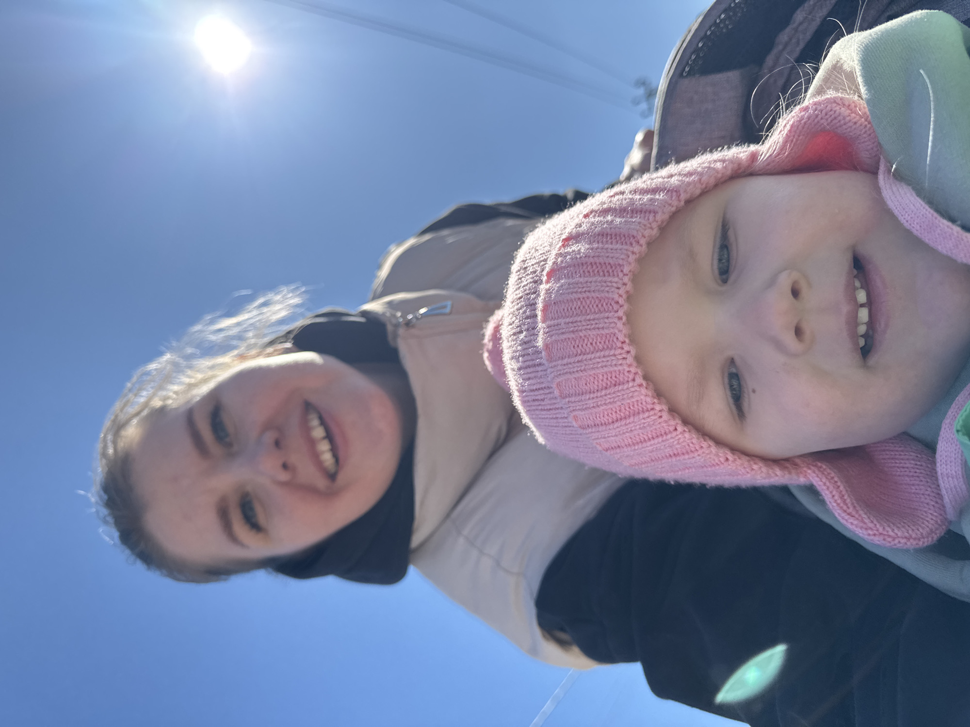 The image depicts a selfie of a woman and a young child taken outdoors on a bright, sunny day. The woman, with fair skin and light hair, is smiling and wearing a black jacket with a beige lining. The child, who is positioned closer to the camera, is wearing a pink knitted hat. Both subjects are facing the camera.
The background consists of a clear blue sky with the sun visible at the top of the frame. Power lines are also visible in the background. The location appears to be Горный, Russia.