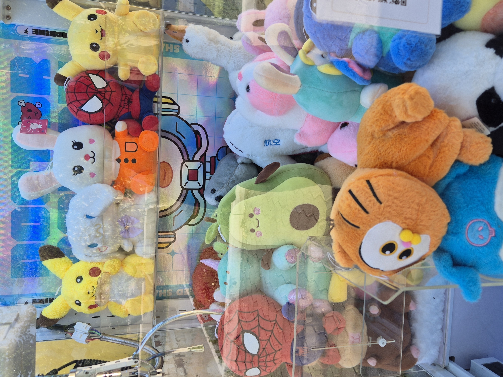 The image shows a claw machine filled with stuffed toys. The toys include Pikachu, Spiderman, a rabbit, a goose, an avocado, and other assorted plushies. The machine is located indoors, the location is unknown.