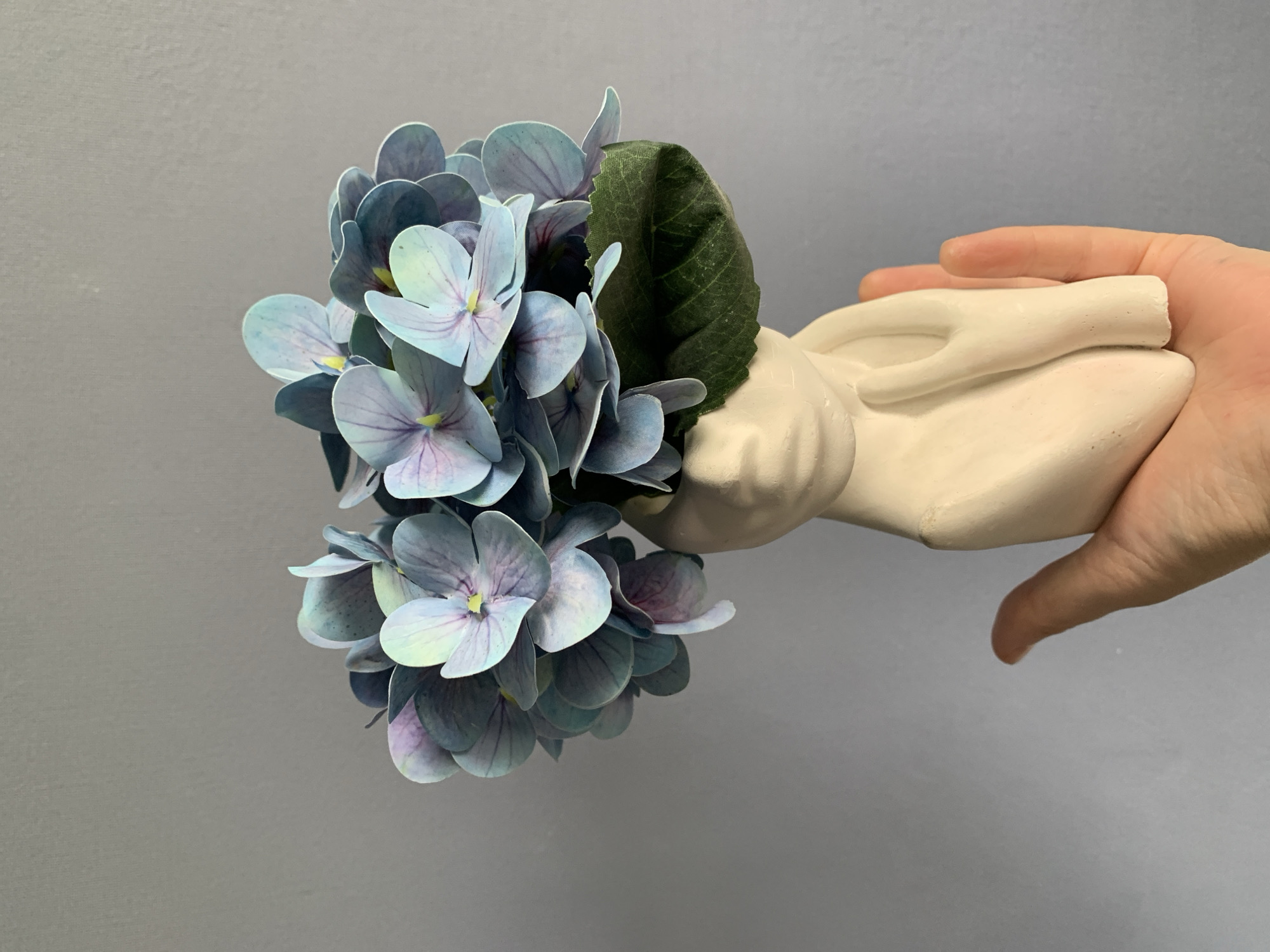A close-up shot shows a white, ceramic hand-shaped vase being held in a human hand against a plain gray background. The vase is filled with a bouquet of blue-purple hydrangea flowers, along with one green leaf. The setting does not depict a city.