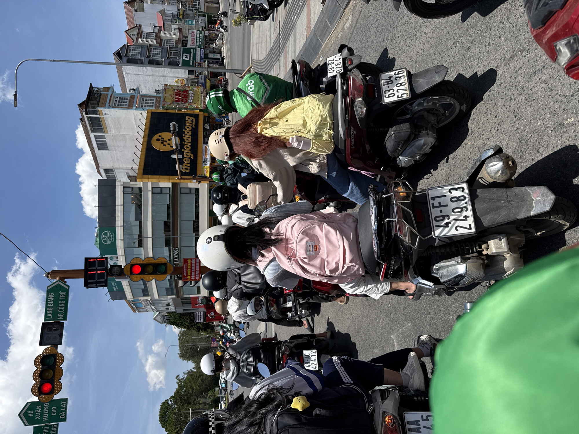 The image captures a bustling street scene in Da Lat, Vietnam, where a multitude of scooters are stopped at a traffic light. Red lights are illuminated on the traffic signals. Signage indicates the location as near Ho Xuan Huong and the road Lang Biang. A large "thegioididong" sign is visible. The scooters' license plates are visible, including examples such as 49-F1 249.83, 63-B4 528.36, 49-82 026.42, and AA 45. Many scooter riders are wearing helmets. Some individuals in green uniforms and "Grab" bags are present. The surrounding buildings and shops are visible in the background under a clear sky.