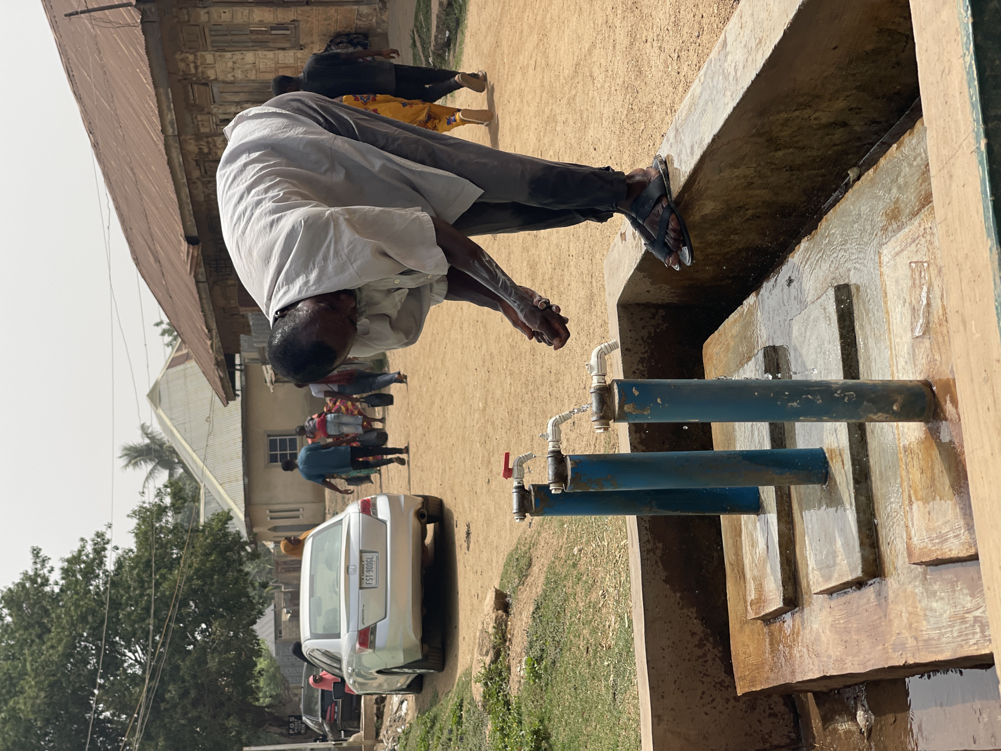 The image depicts an outdoor scene in Muchiya, Nigeria, on what appears to be a bright, sunny day. The primary subject is a man, dressed in a white long-sleeved shirt and dark trousers, bending over a concrete basin to wash his hands and face. His hands are cupped under a stream of water, and his face is lowered towards the basin. He is wearing black sandals, with one foot resting on the edge of the basin.
In the immediate foreground, a series of public water taps are visible, constructed with two blue vertical pipes supporting white PVC piping and red shut-off valves. Water is actively flowing from at least one tap into the rectangular concrete basin below. The ground surrounding the water point is sandy and dusty.
In the background, several other individuals are present, some walking and others standing, though their faces and details are blurred. A silver Honda Accord sedan is parked further back, displaying the license plate "FST 900GL," typical of Nigerian registration. A small, partially visible sticker on the car's rear might read "PDS." Beyond the car and people, various buildings with light-colored walls and roofs, along with green trees, line the dusty street, indicating a community setting. The bright lighting suggests it is daytime.