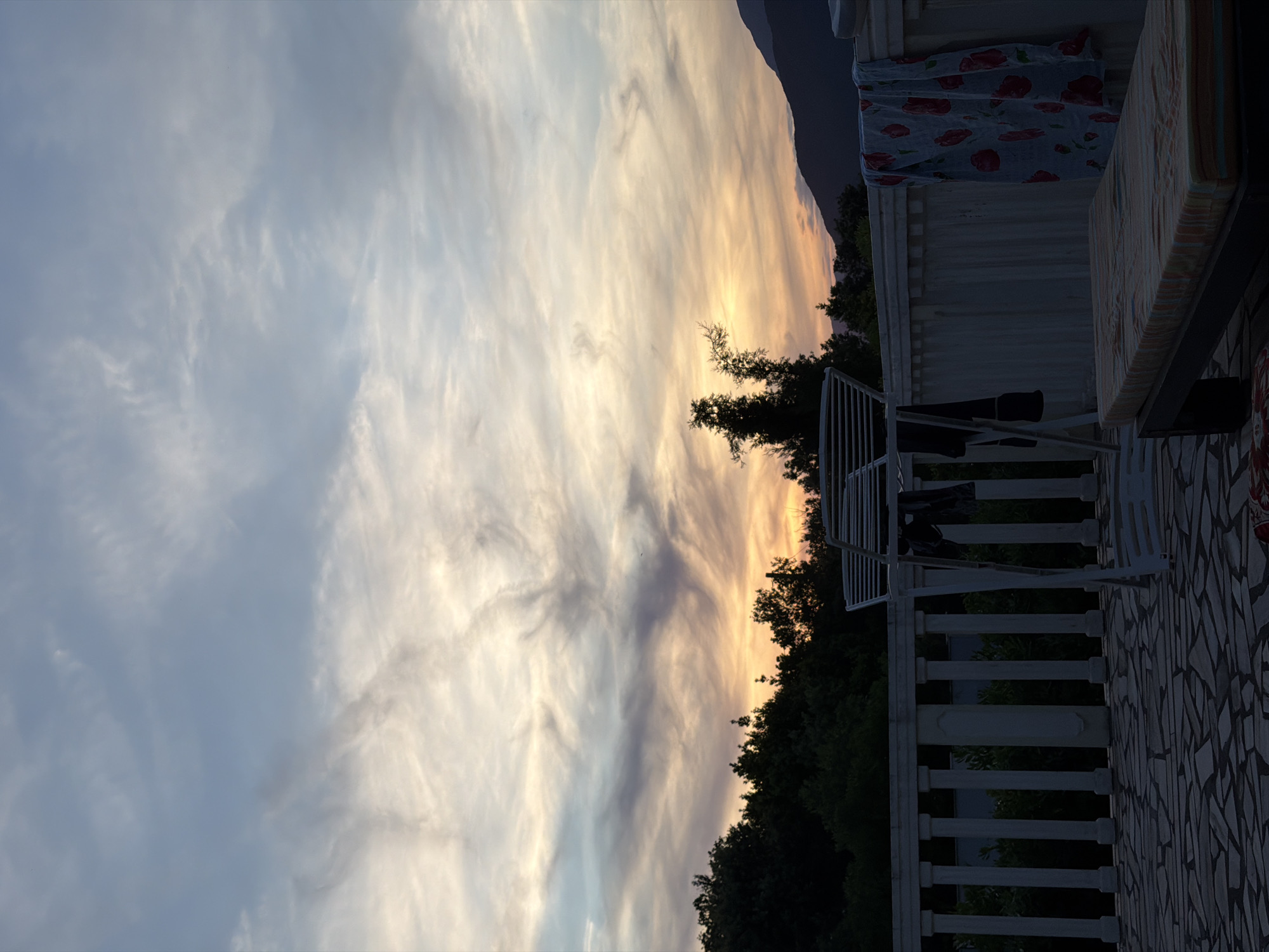 The image shows an outdoor scene in Gošići, Montenegro, at dusk or dawn. The sky is filled with clouds, ranging from light gray to yellow and orange near the horizon, indicating sunrise or sunset. Silhouetted trees and possibly a mountain are visible against the vibrant sky. A building or structure with a mosaic tile floor and a white railing is in the foreground. A clothes drying rack stands on the terrace. A patterned fabric is draped over the side of the building.