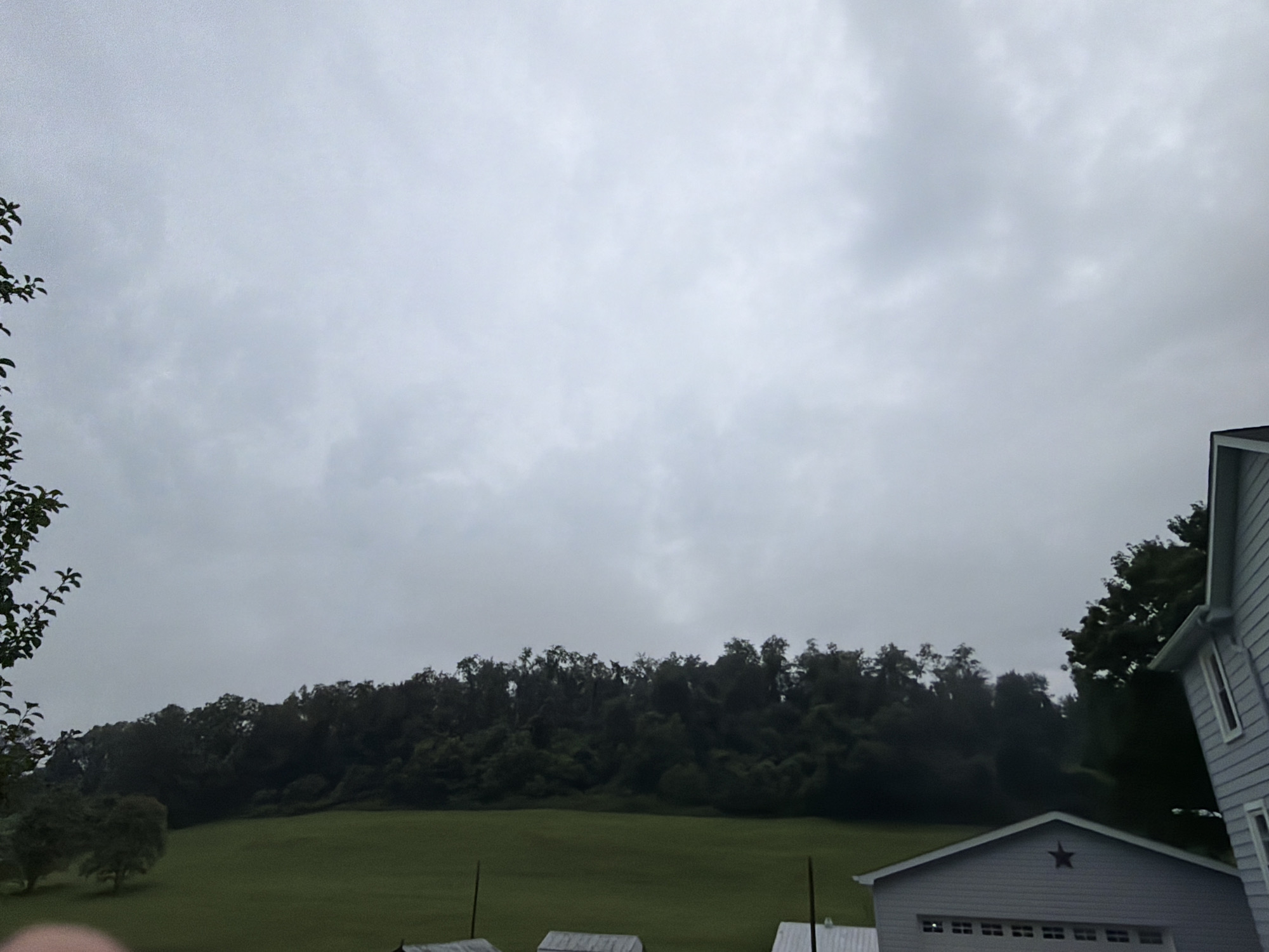 The image shows a rural landscape under an overcast sky. In the foreground, there is a gray building with a garage door that has a star-shaped decoration above it. To the right of this building, a portion of a light blue house is visible, showing its siding and a window. A grassy field stretches out from the buildings towards a dense line of dark green trees that form a forest along a hillside. The trees are lush and full, suggesting a temperate climate. The sky is a uniform light gray, indicating cloudy weather, possibly pre-storm conditions. There are no people clearly visible in the scene. The location is identified as Fallowfield Township, United States of America.