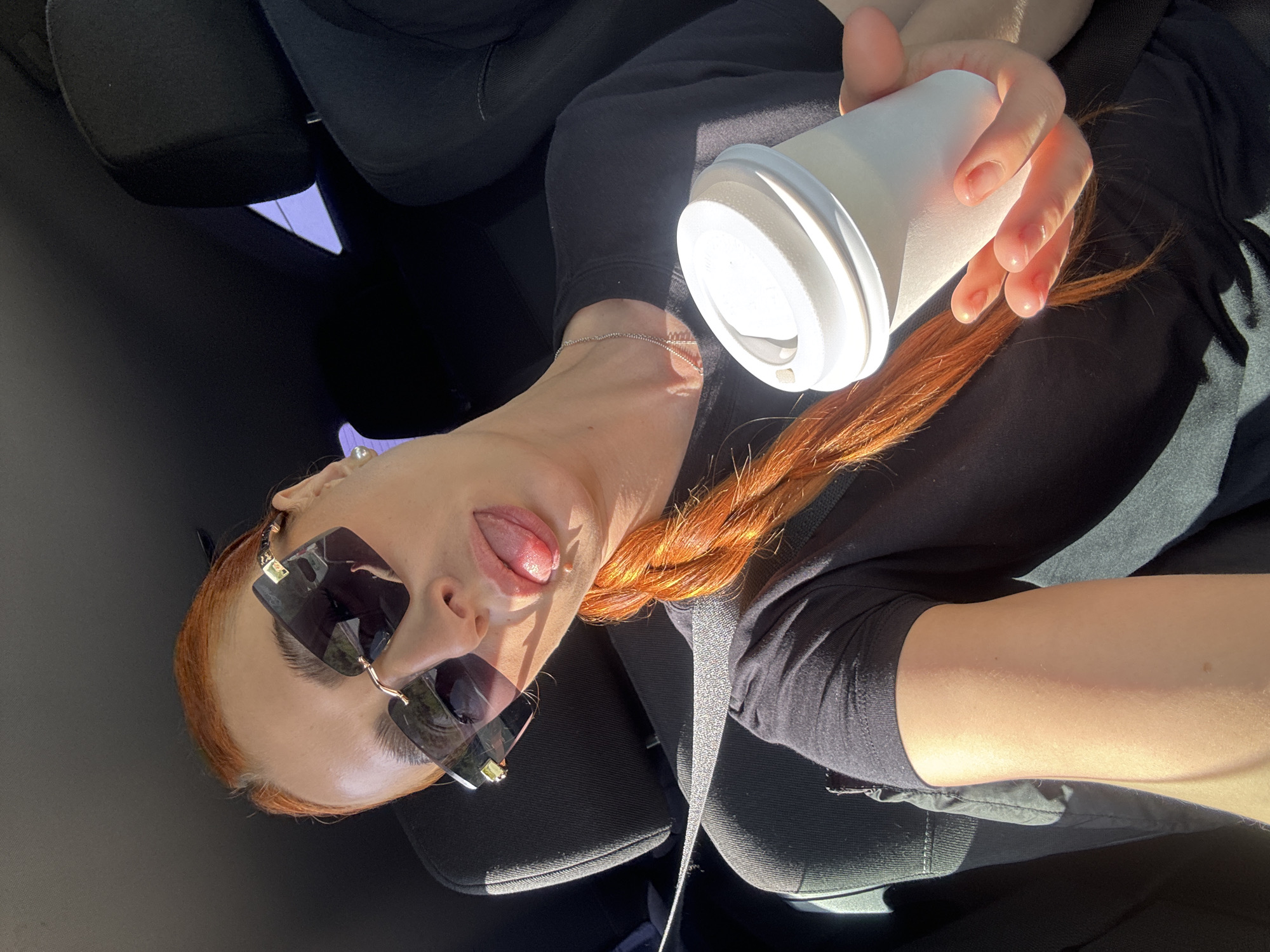 A woman with red braided hair and sunglasses is sitting in a car, holding a white paper cup. She is wearing a black long-sleeved shirt. The interior of the car is black. The car's seatbelt is visible. The woman is sticking her tongue out. The location is unlikely to be identified as Podlastva (Poljice), Montenegro from just the scene description.