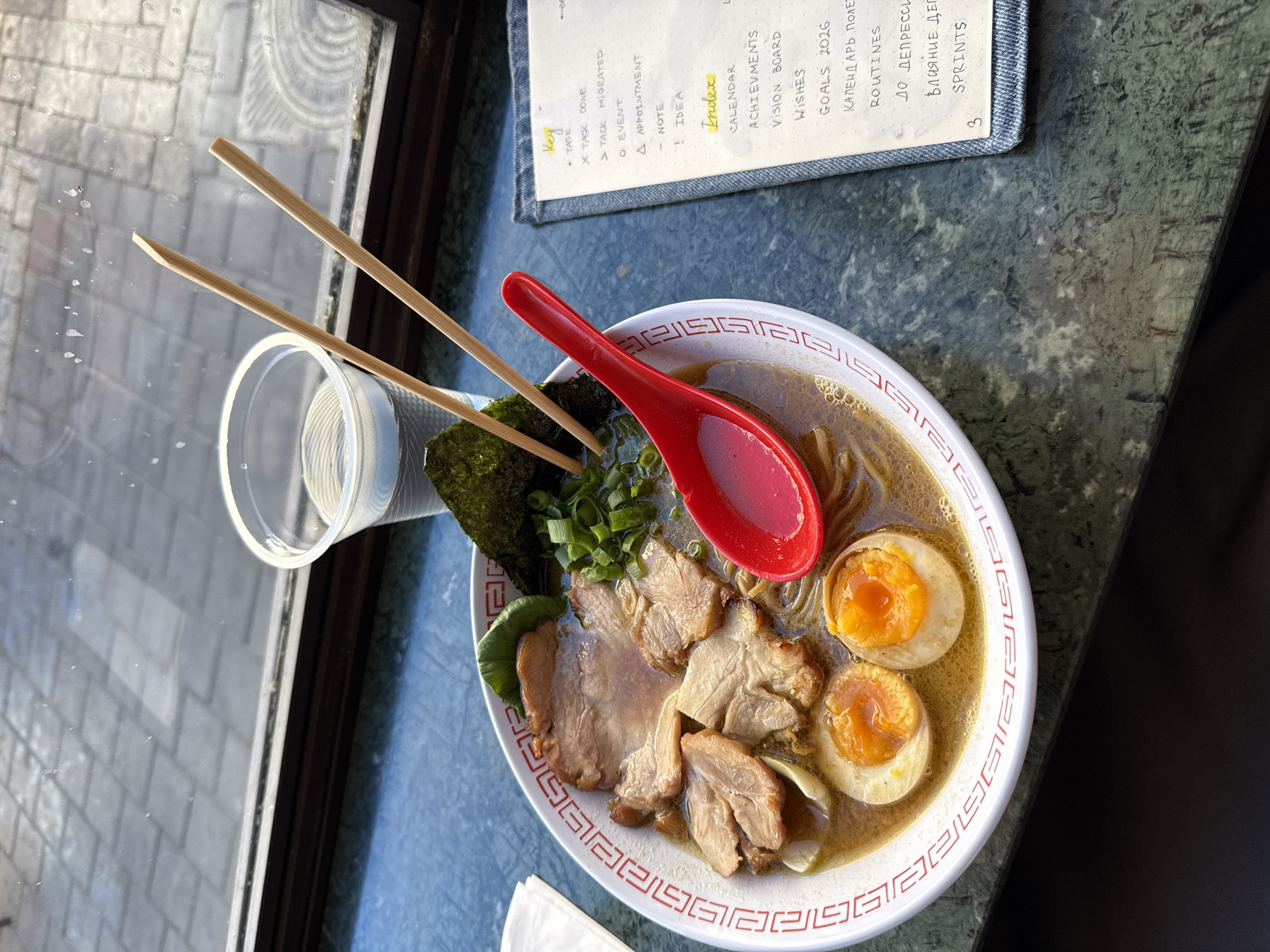 A bowl of ramen sits on a green marble countertop next to a plastic cup of water and a piece of paper with a handwritten to-do list. The ramen is topped with slices of pork, green onions, nori seaweed, and two soft-boiled eggs. Chopsticks rest on top of the bowl, and a red ladle is placed on the side. The to-do list includes sections for "Key," "Index," and various tasks and goals, with some entries in Russian. In the background, a window shows a view of a cobblestone street. The lighting suggests it is daytime.
