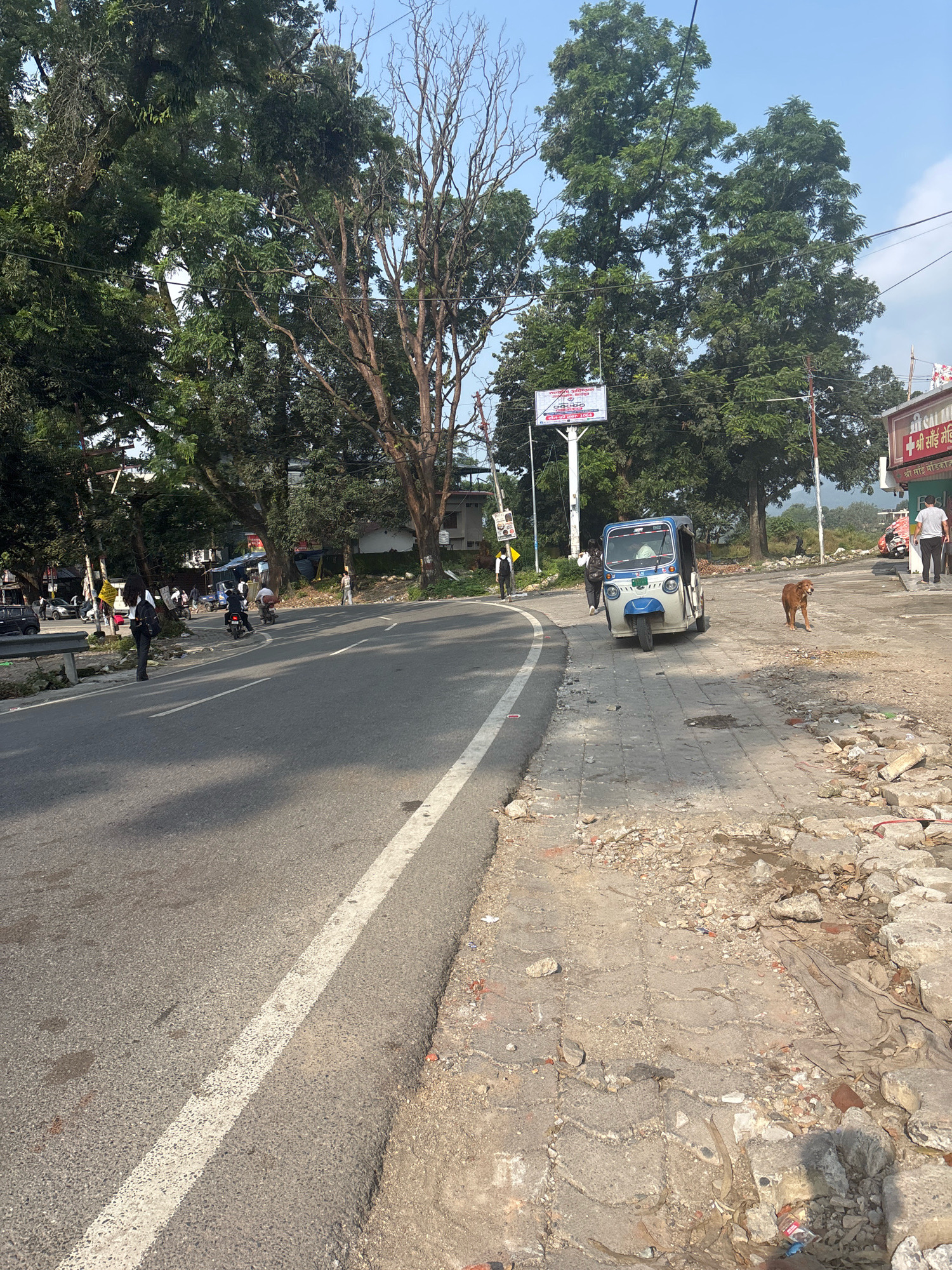 The image depicts a street scene in Bhatta, India, on a sunny day. A two-lane asphalt road occupies the foreground, with a white line dividing the lanes. The road curves to the left, leading into the distance. On the right side of the road, a concrete sidewalk is visible, with piles of rubble and debris suggesting construction or demolition. A brown dog stands on the sidewalk, facing left.
Further down the road, an auto-rickshaw, predominantly white with blue accents, is parked on the right side. Several people are seen walking or standing near the auto-rickshaw and on the sidewalk. A woman in dark clothing with a backpack is walking away from the auto-rickshaw towards the left side of the road. Other individuals are blurred in the background.
The left side of the image features a shoulder with a metal guardrail and more rubble. Lush green trees with dense foliage line the background, interspersed with some sparser, bare trees. A few buildings and structures are visible behind the trees, along with utility poles and wires. Two signs are mounted on poles: one on the left side with text in Hindi, and another on the right displaying the "Shri Sai Medical" logo. The sky is clear and blue.