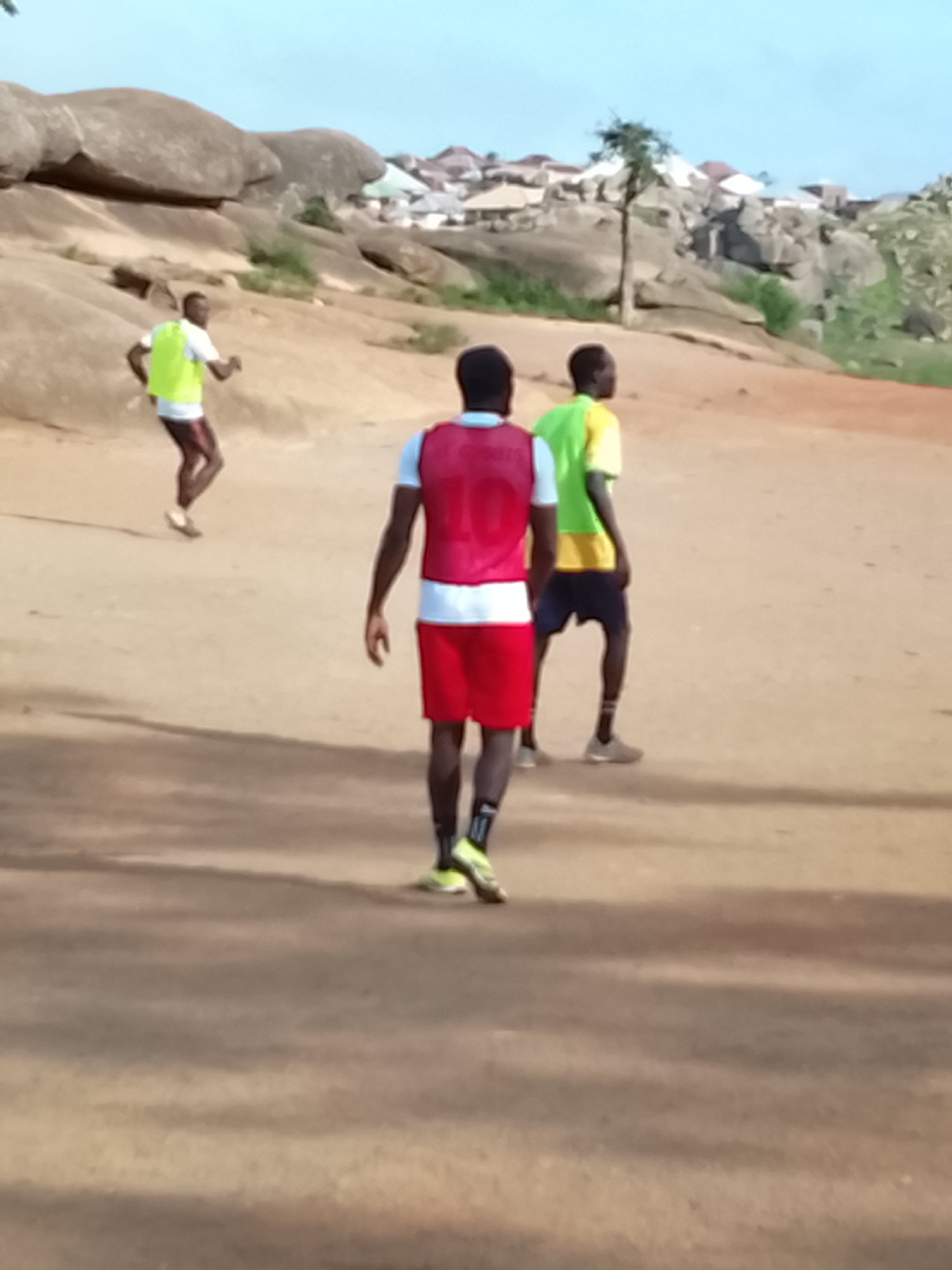 Three individuals, appearing to be young men, are engaged in an outdoor activity on a dusty, uneven terrain, possibly a training ground. In the foreground, one person faces away from the camera, wearing a red vest over a white t-shirt and red shorts. He has dark skin and dark hair, and is wearing dark socks and bright yellow athletic shoes. To his left and further back, another individual is also facing away, wearing a bright yellow and green t-shirt, dark shorts, and grey athletic shoes. Further to the left and in the background, a third person, also appearing to be a young man, is in mid-stride, running away from the camera. He is wearing a bright yellow vest over a white t-shirt and dark shorts, with dark skin and dark hair.
The environment is characterized by a dry, sandy ground. In the background, large, weathered rock formations dominate the landscape, with a scattering of buildings and vegetation visible on and around them, suggesting an elevated or hilly urban fringe. The sky is clear and light blue. This scene is consistent with locations in the city of Jos, Nigeria, known for its rocky terrain and outdoor recreational spaces. The presence of athletic attire and the running posture suggest a sports-related activity.