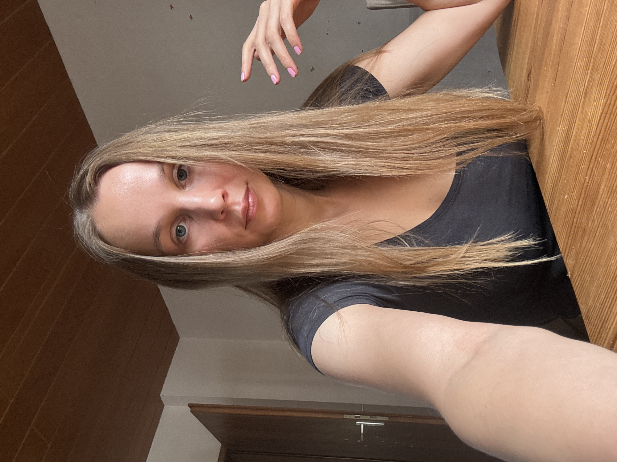 This is a medium-shot, indoor, selfie-style photograph of a Caucasian woman with long, blonde hair. She is wearing a dark grey t-shirt. Her left arm is extended towards the camera, and her right hand is resting on the table in front of her, with painted pink fingernails. She has light blue eyes and is looking directly at the camera. The background shows a wooden ceiling, a white wall with some small, red-colored marks, and a wooden door with a silver handle on the left side of the frame. The woman is seated at a wooden table. The scene appears to be inside a residential setting, possibly in Überherrn, Germany, given the architectural style of the ceiling and door.