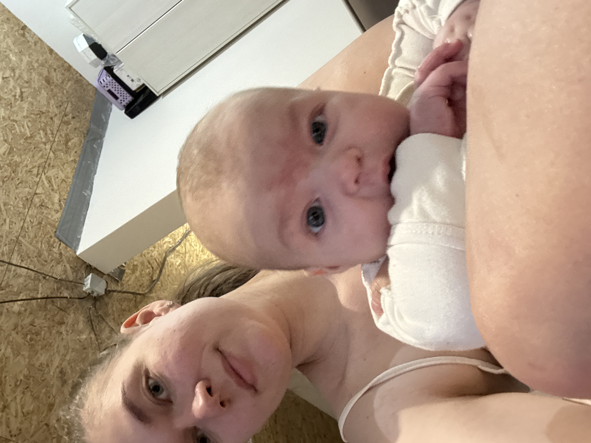 A low-angle selfie taken indoors shows a woman and a baby, likely in Chaikovsky, Russia. The baby, fair-skinned with light hair and blue eyes, is in the foreground, looking directly at the camera. The baby is wearing a white garment. The woman, fair-skinned with light brown hair, is visible in the lower left corner, also looking at the camera. The background consists of light-colored walls, a white doorframe, and electrical wiring.