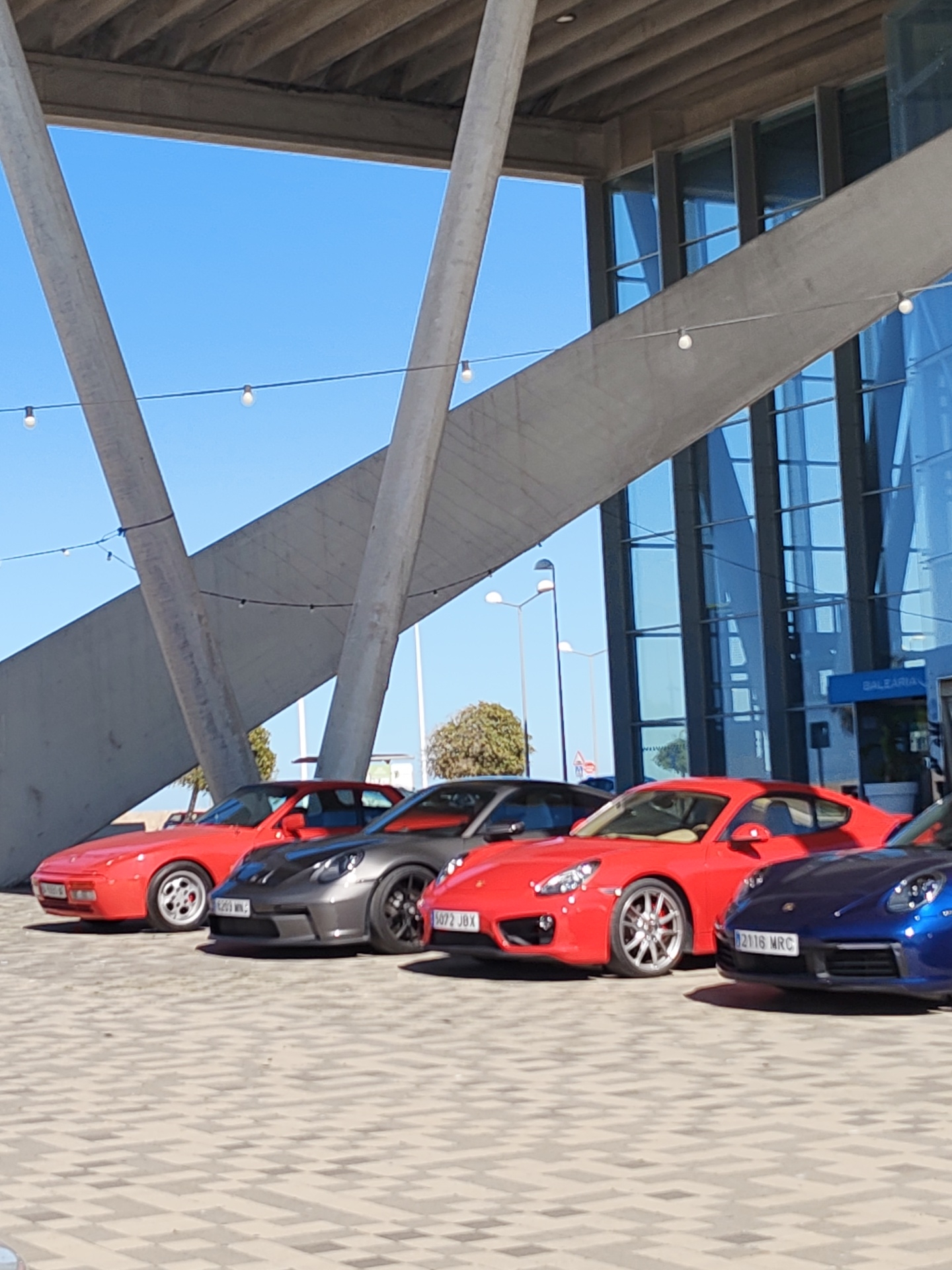 The image shows a row of five Porsche cars parked in front of a modern building with glass windows and a striking concrete structural design. The cars, from left to right, are red, gray, red, red, and blue. The building has a sign that reads "Baleària." The location is Dénia, Spain, based on the presence of the Baleària terminal. The cars are parked on a patterned paved surface. The sky is blue and clear. Overhead lighting is visible, strung on wires.