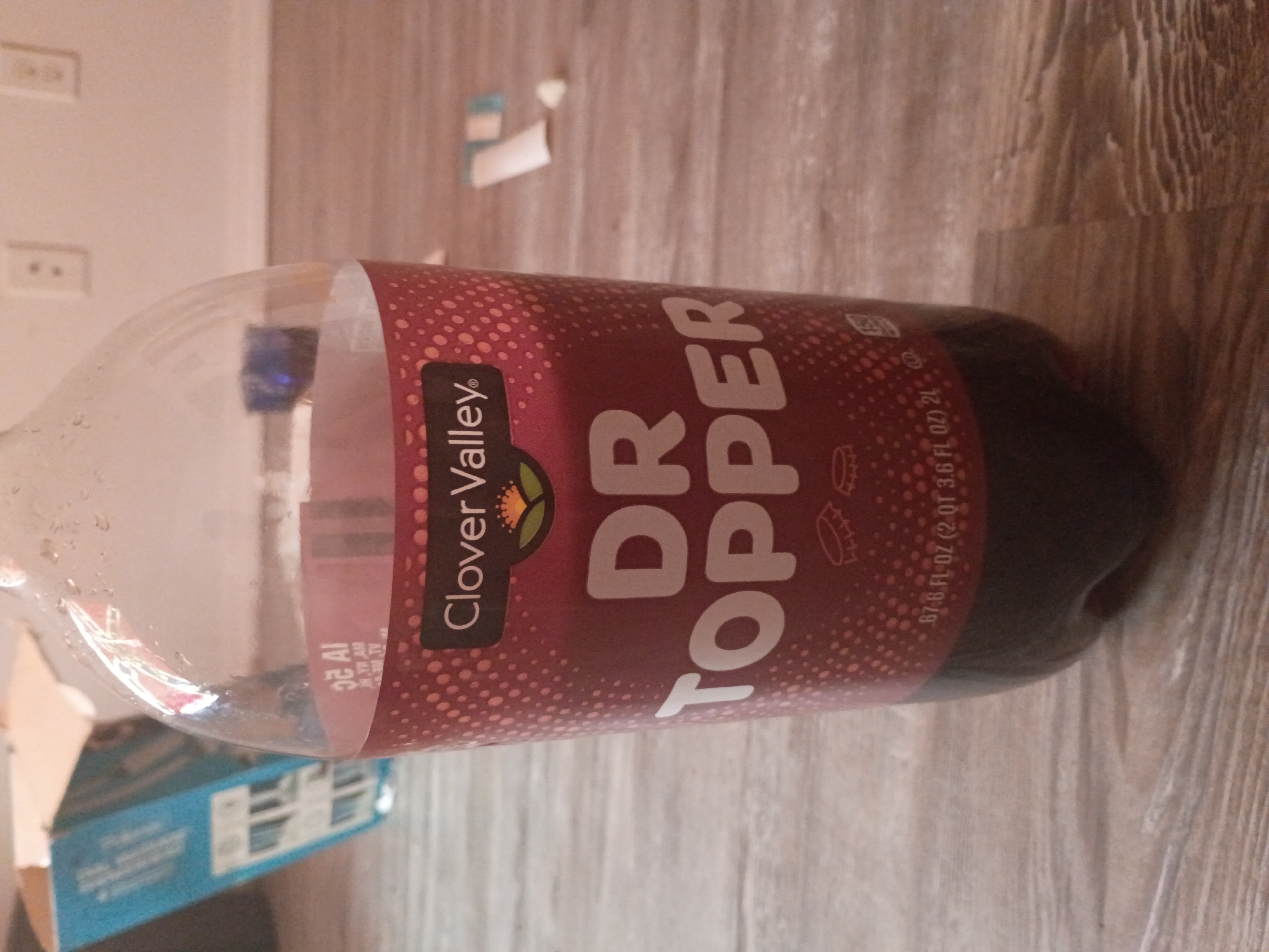 A nearly empty 2-liter bottle of "Dr. Topper" soda, a Clover Valley brand, lies horizontally on a light-colored wooden floor. The bottle is positioned so that the label is facing the viewer. Part of a blue box is visible in the lower left corner of the image. The background also includes a white wall with electrical outlets. The setting is indoors, likely in a home, but there is no indication of a location in Clarksville, United States of America.
