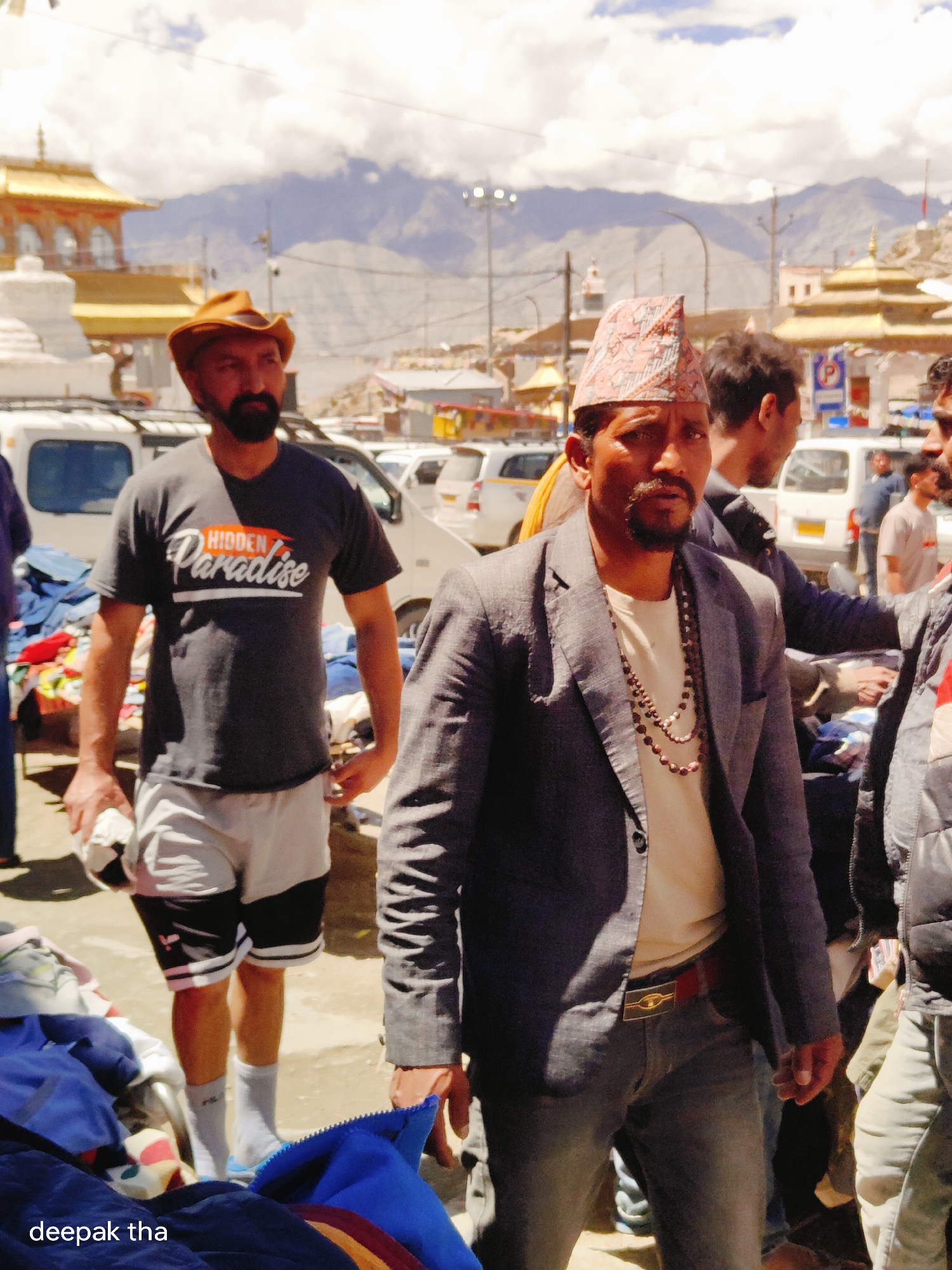 The image depicts a street scene in the city of Leh, India, likely in the vicinity of a market or public area given the presence of stalls and various vehicles. The backdrop features traditional Ladakhi architecture with golden roofs and stupas, set against a mountainous landscape under a cloudy sky.
In the foreground, three men are the primary subjects. The man on the left is wearing a dark grey t-shirt with "HIDDEN Paradise" written on it, light grey shorts with black accents, and a brown cowboy hat. He has a beard and appears to be holding a white object. To his right, a man in a grey blazer, a light-colored t-shirt, and a traditional patterned cap is looking directly at the camera. He has a beard and is wearing a beaded necklace. The man on the far right is partially visible, wearing a dark jacket.
Several white vehicles, likely minivans or SUVs, are parked in the background, suggesting a busy thoroughfare or parking area. Stalls with various goods, including clothing, are visible, indicating a market environment. The overall lighting suggests it is daytime. The text "deepak tha" is watermarked on the bottom left of the image.
