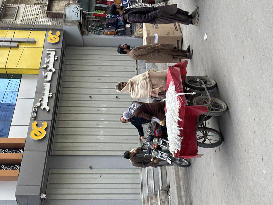 The image depicts a street scene in Quetta City Tehsil, Pakistan, during daylight hours.
In the immediate foreground, a red four-wheeled pushcart is stationary on an asphalt road. The cart is covered with a red tarp and laden with numerous small, white, fibrous, cylindrical, or string-like items.
In the midground, six adult males are present. From left to right:
1. A male in a brown jacket and light-colored scarf or shawl around his neck stands next to a parked silver and red motorcycle, looking downwards.
2. A male in a dark shirt is positioned behind the motorcycle, facing another individual.
3. A male in a brown jacket/shirt, with his head covered by a light-colored shawl or scarf, stands next to the motorcycle, holding a yellow object and facing away, appearing to interact with the individual behind the motorcycle.
4. A male in light brown traditional attire (shalwar kameez) with a large, light-colored shawl draped over his head and shoulders stands facing forward, slightly to the left.
5. A male in brown traditional attire (shalwar kameez) stands with hands by his sides, facing left.
6. A male in a dark jacket/coat and dark traditional trousers stands with hands in his pockets, facing left, near a large, upright brown cardboard box with partially visible "Haier" branding on the right sidewalk.
The background features commercial buildings. The prominent structure directly behind the individuals has a grey facade with closed corrugated metal shutters. Above the shutters, a sign reads "OC الیکٹرک سینٹر" (OC Electric Center) in Urdu script, flanked by "OC" logos. Higher sections of this building feature yellow and blue elements. To the far right, another storefront with various items, possibly bags and garments, displayed outside is partially visible, with a small sign containing partial Urdu text, "بازار نگ." The road surface is grey asphalt, and a concrete curb and steps are visible on the left.
