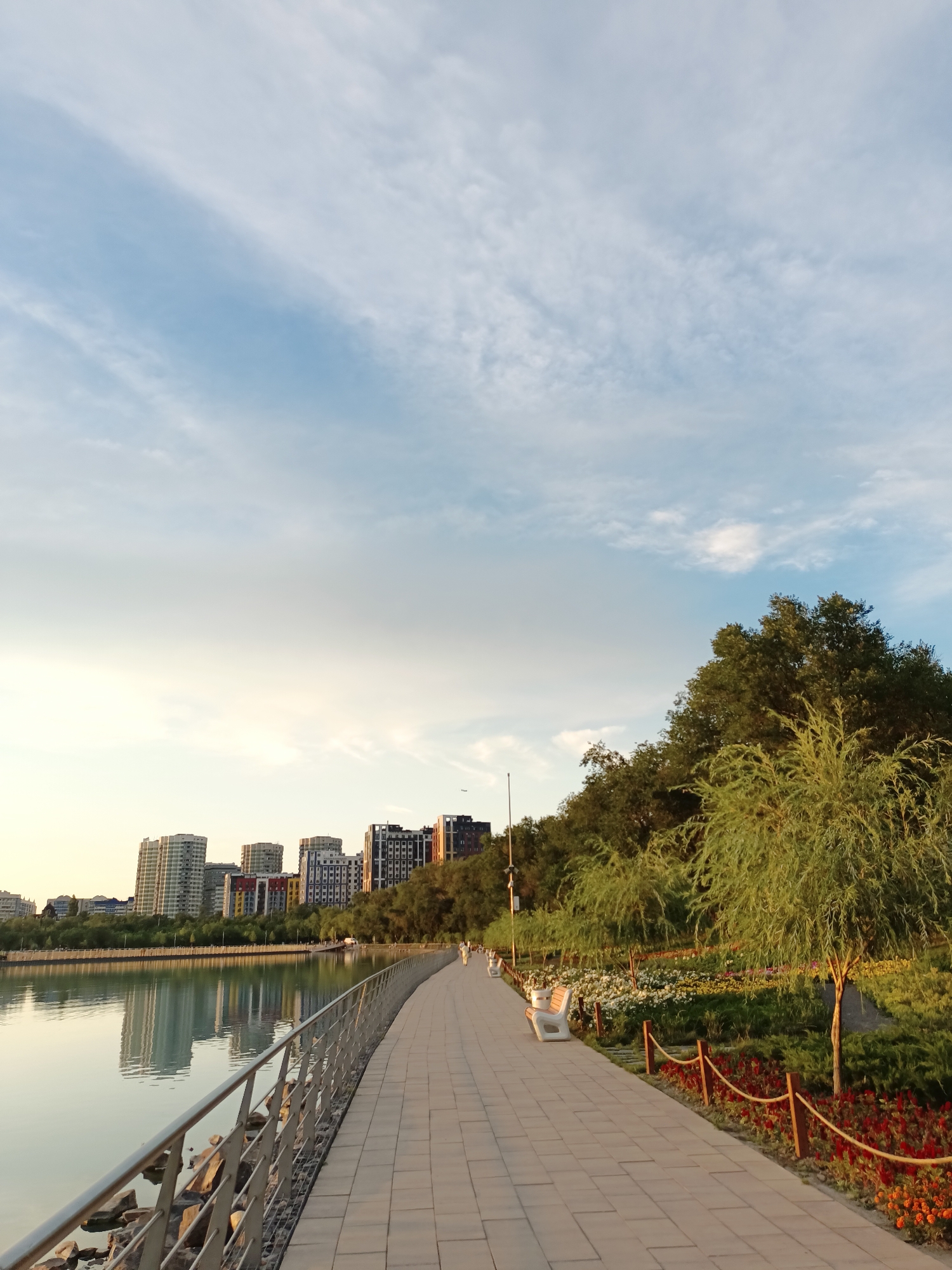 The image depicts a scenic waterfront pathway in Almaty, Kazakhstan, likely within the city's urban parks or near a body of water like a lake or river. The scene is captured during what appears to be late afternoon or early evening, with soft, warm sunlight illuminating the landscape and casting gentle reflections on the water.
In the foreground, a paved walkway stretches along the edge of a calm body of water. A silver metal railing lines the left side of the path, separating it from the water's edge where rocks are visible. The walkway is made of rectangular pavers laid in a uniform pattern. Several white park benches are placed along the right side of the pathway, offering spots for rest and contemplation.
Adjacent to the walkway on the right is a beautifully landscaped garden area. It features a variety of flowers in vibrant colors, including red and orange, interspersed with lush green bushes and manicured trees. A rope barrier with wooden posts delineates the garden from the walkway. Further in the background, a line of trees forms a natural backdrop to the garden, with their leaves catching the golden light.
Across the water, a cityscape of modern buildings is visible. These high-rise structures, varying in height and architectural style, are reflected in the tranquil water. Some buildings are modern glass and steel designs, while others have colorful facades. The reflection creates a mirrored image of the urban skyline on the water's surface.
The sky above is a clear blue, with wispy white clouds scattered across it, suggesting pleasant weather. The overall atmosphere of the image is peaceful and serene, inviting viewers to imagine a leisurely stroll or a moment of relaxation in a picturesque urban setting.