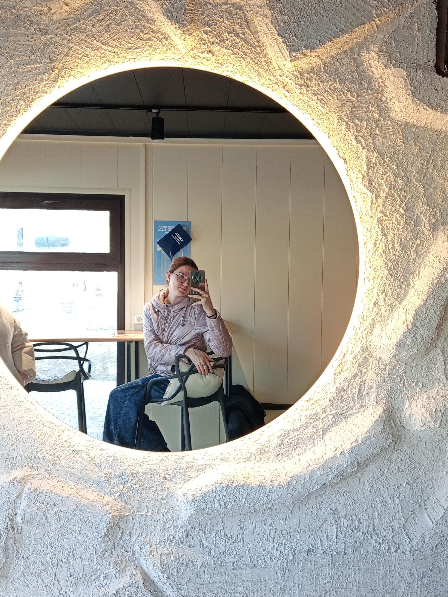 The image displays a mirror selfie taken in an indoor setting within the city of Chaikovsky, Russia. A female individual with red hair and glasses, dressed in a light pink puffer jacket and blue jeans, is seated in a chair and holds a smartphone with a green case to capture her reflection. She exhibits a slight smile.
The mirror itself is large and circular, framed by a thick, white, highly textured material resembling rough plaster with undulating forms. The inner edge of the frame emits a soft, integrated light, creating a luminous halo around the reflection.
Within the mirror's reflection, the individual is positioned against a light beige, vertically paneled wall. To her left, a large window with a dark brown frame provides a bright, blurred view of an outdoor environment. A narrow wooden counter extends beneath the window, and a second, similar chair is partially visible to the far left. Above the reflected figure, a black track light fixture is mounted on a dark ceiling panel. A small, rectangular blue sign with white, mirrored text, appearing as "ОТЕЛ" on its top line, is visible on the wall to the right of the individual. The scene suggests a cafe or a lounge area.