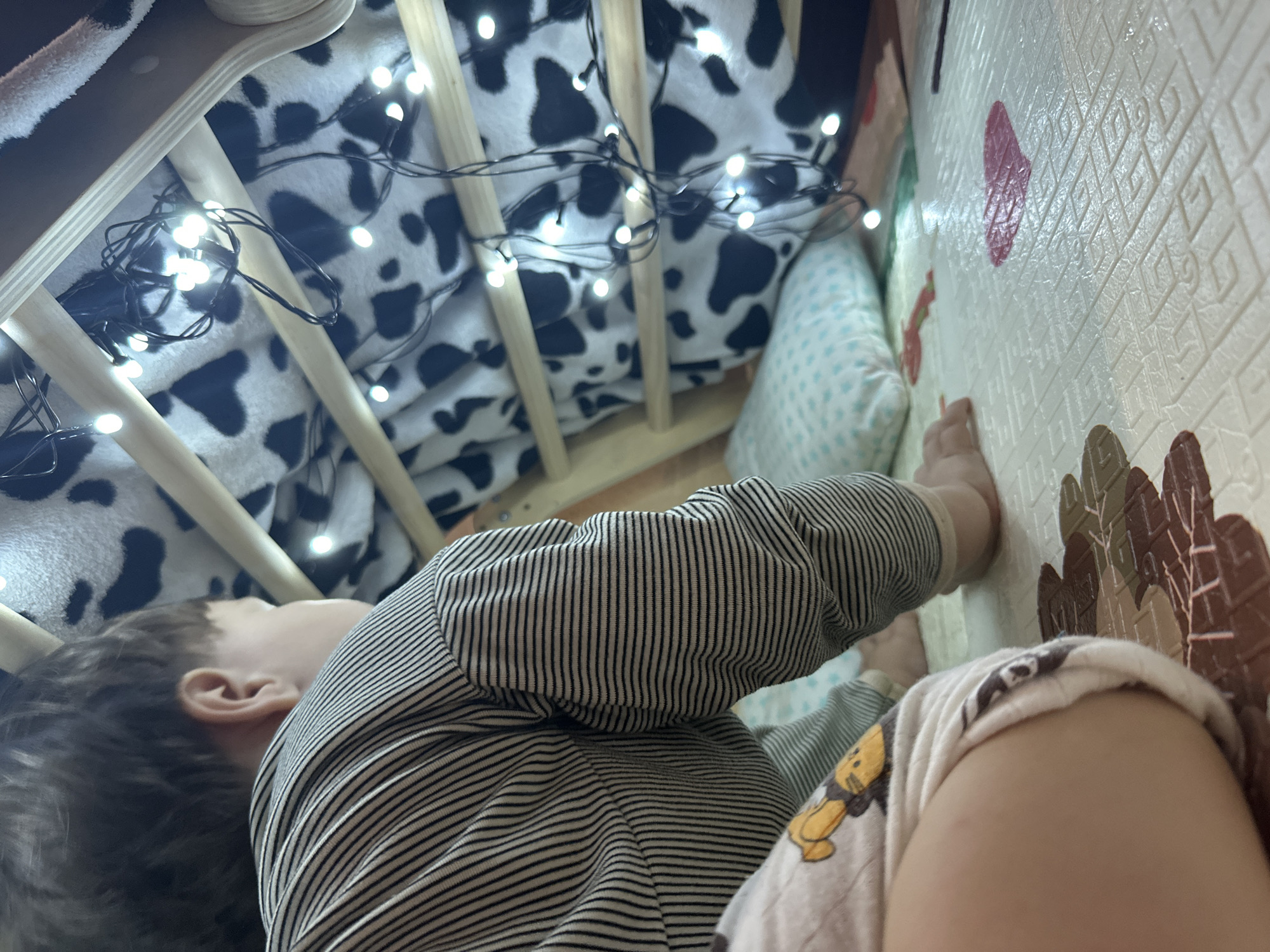 A child is inside a tent structure made of wooden beams and covered with a black and white cow-print fabric. White string lights are draped around the inside of the tent, illuminating the space. The child, wearing a striped shirt and patterned shorts, is on hands and knees on a light-colored playmat with embossed geometric patterns and some colorful shapes, including a red heart. A light blue pillow with white dots is visible behind the child. The scene appears to be indoors. The location is not specified in the image.