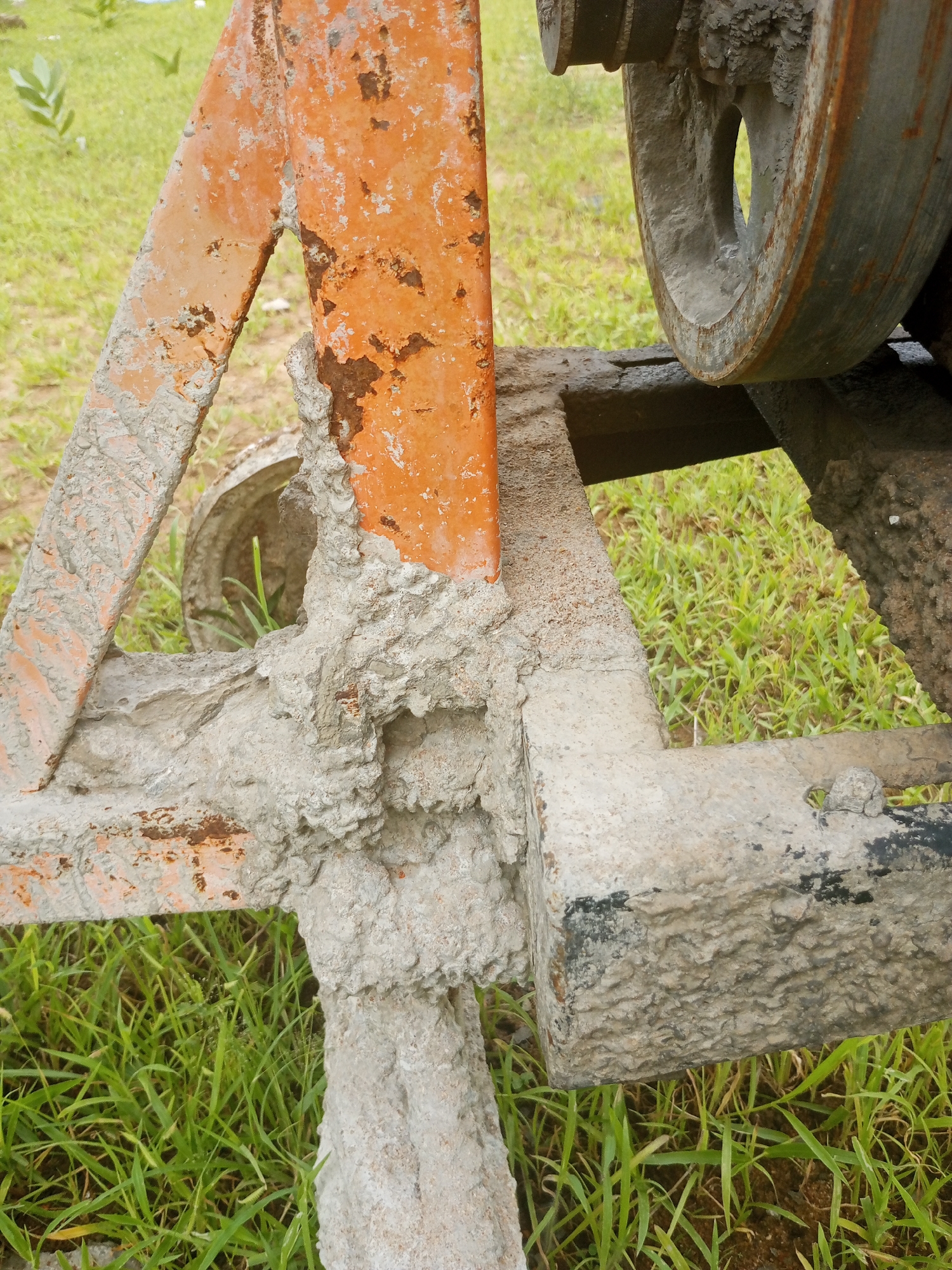 The image displays a close-up view of a concrete mixer, specifically its frame and a wheel. The metal frame, which appears to be painted orange, is heavily corroded with rust and has dried concrete caked onto it in several places. One of the mixer's wheels is visible, also partially covered in dried concrete. The background consists of green grass and some soil, suggesting an outdoor setting. The lighting appears to be natural daylight, with no strong shadows or indications of a specific time of day. No people are visible in the image, and no text is present. The scene conveys a sense of disuse or post-construction activity, given the state of the equipment and the presence of dried concrete. The location context of Jalingo, Nigeria, places this scene in a tropical environment, which could explain the lush green grass.