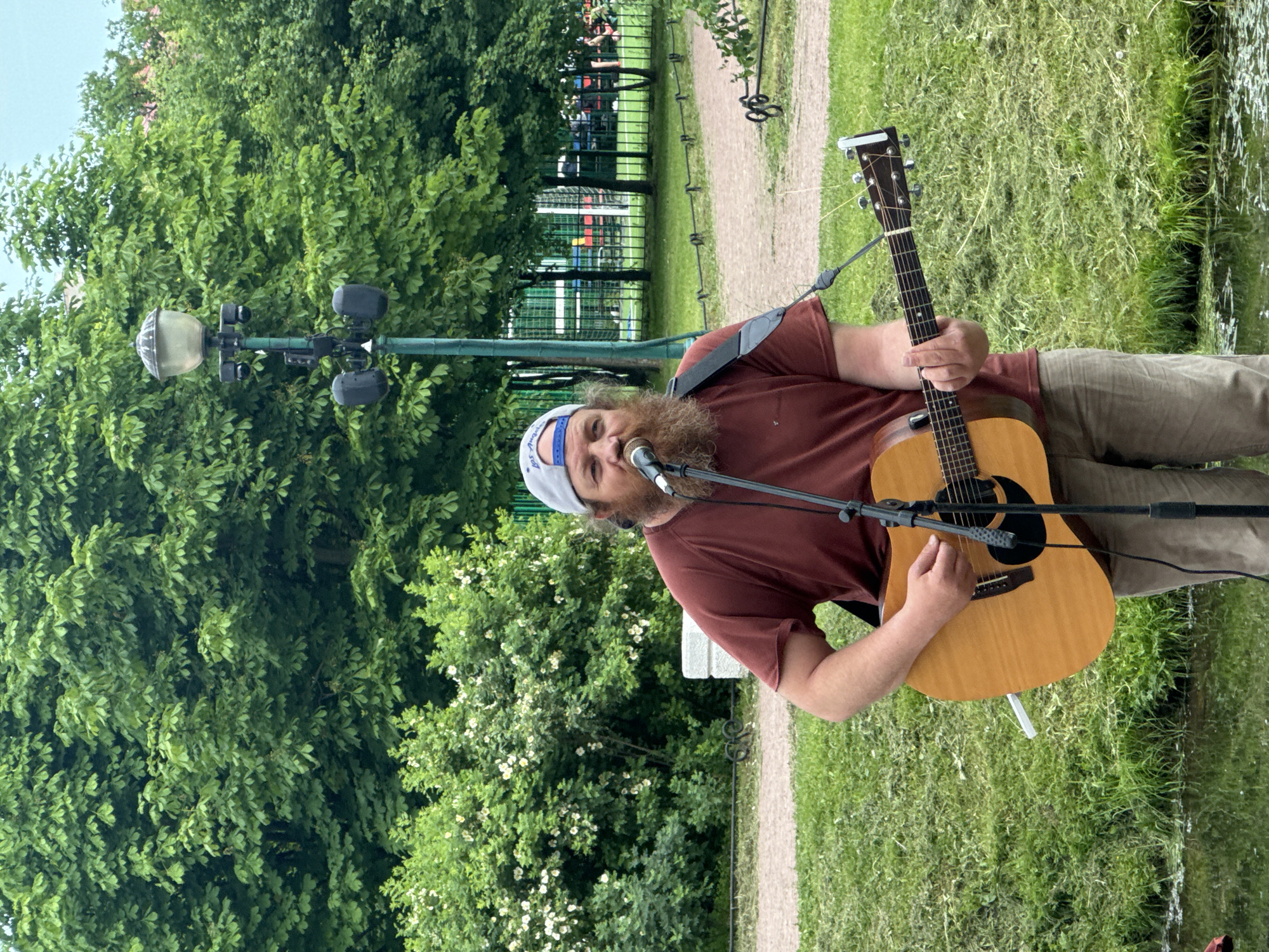 A man with a beard, wearing a maroon t-shirt and a grey baseball cap, plays an acoustic guitar in a park. He is standing in front of a microphone with his guitar strap over his right shoulder. Behind him, there are lush green trees and bushes, with a lamppost and speakers in the background. A body of water is visible in the foreground. The location appears to be a park in Saint Petersburg, Russia, possibly near a sports field.