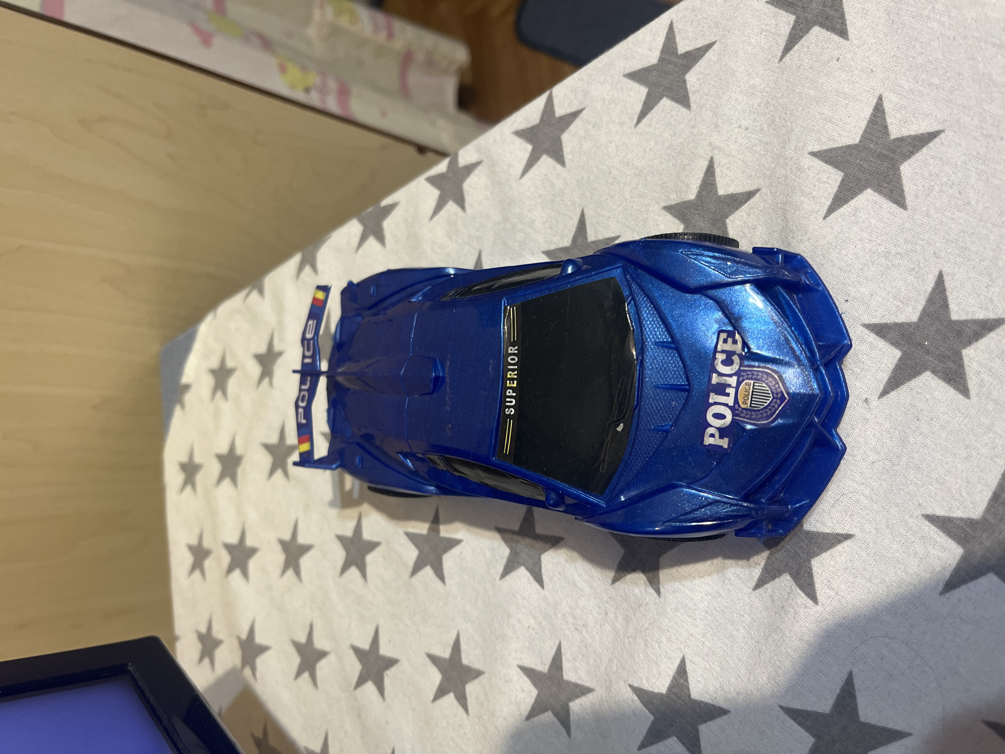 A metallic blue toy police car with a futuristic design is the central subject, positioned on a white fabric surface patterned with numerous dark grey five-pointed stars. The car's windshield bears the word "SUPERIOR" in gold lettering, accompanied by three parallel gold lines above it. The front hood displays "POLICE" in white, situated above a purple and silver shield-like emblem also reading "POLICE." The rear spoiler likewise features "POLICE" in white, beneath a small vertical tri-stripe of red, yellow, and blue. The toy appears to be in good condition.
To the left of the main subject, a portion of a black television or monitor frame is visible, showing a blue display. The background consists primarily of light-colored wooden paneling or furniture. On the far right, a vertical section of a faintly patterned, light-toned fabric, possibly a curtain, is partially visible. The scene is illuminated from the upper left, casting distinct shadows from the toy car onto the star-patterned fabric. The environment is distinctly indoors. No specific visual cues within the image definitively confirm its location within the city of Burgas, Bulgaria.