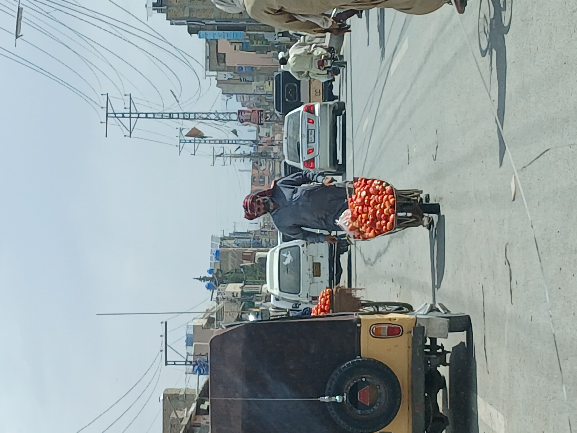 The scene shows a street in Quetta City Tehsil, Pakistan. A man wearing a red head covering is walking along the street, pushing a bicycle with a large container of tomatoes attached. The street has a busy atmosphere with vehicular traffic, including a yellow and black rickshaw in the foreground with a spare tire on the back, a white car, a van, and a person on a scooter. The background displays buildings and utility poles with numerous wires overhead. The sky is hazy, suggesting a bright, sunny day. The overall environment is typical of a busy urban setting in Pakistan.