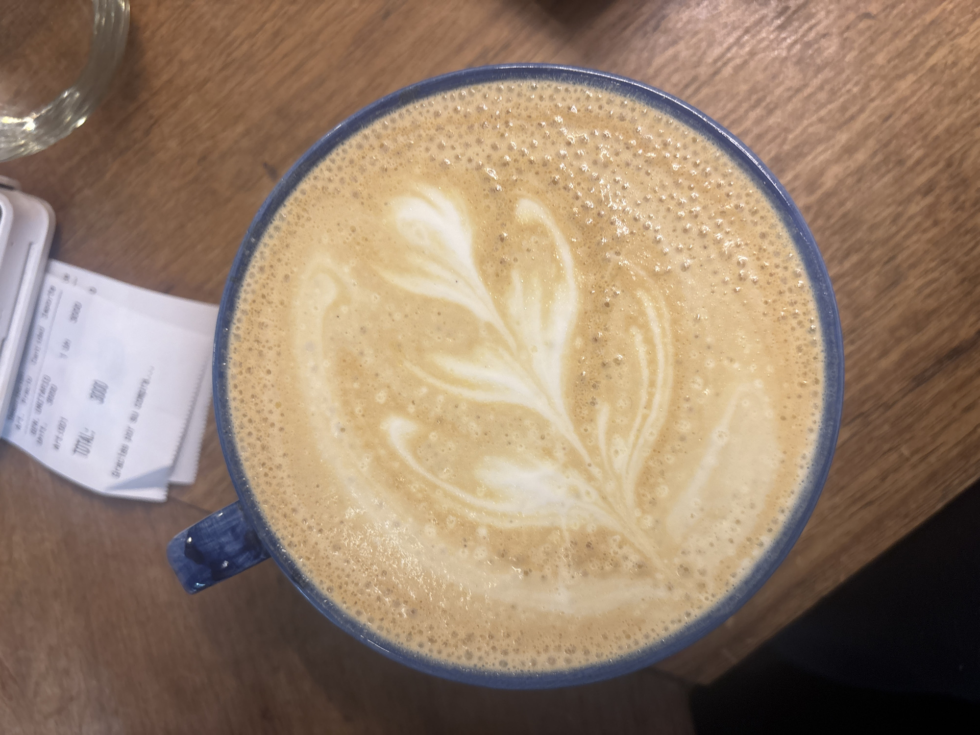 The image presents a top-down view of a cup of coffee, likely a latte, on a wooden table. The prominent subject is the coffee itself, featuring intricate white latte art shaped like a multi-leaf rosetta or floral pattern against the light brown surface of the beverage. The coffee cup is a deep blue or navy color with a slightly textured rim and a handle visible on the left side.
The setting appears to be a cafe or coffee shop in Buenos Aires, Argentina. The table is made of light-colored wood with visible grain. To the upper left of the cup, the rim of a clear glass is partially visible. Further left and slightly below, a paper receipt is crumpled and folded, with some text clearly discernible. The visible text on the receipt includes "TOTAL:", "3000", "1 un", and "Gracias por su compra..." (Spanish for "Thanks for your purchase..."). The "3000" likely represents the cost in Argentine Pesos.
The activity suggested is enjoying a coffee break. The lighting indicates a daytime setting, though the exact time is not specified. The overall impression is one of a relaxed moment, appreciating a well-prepared drink.