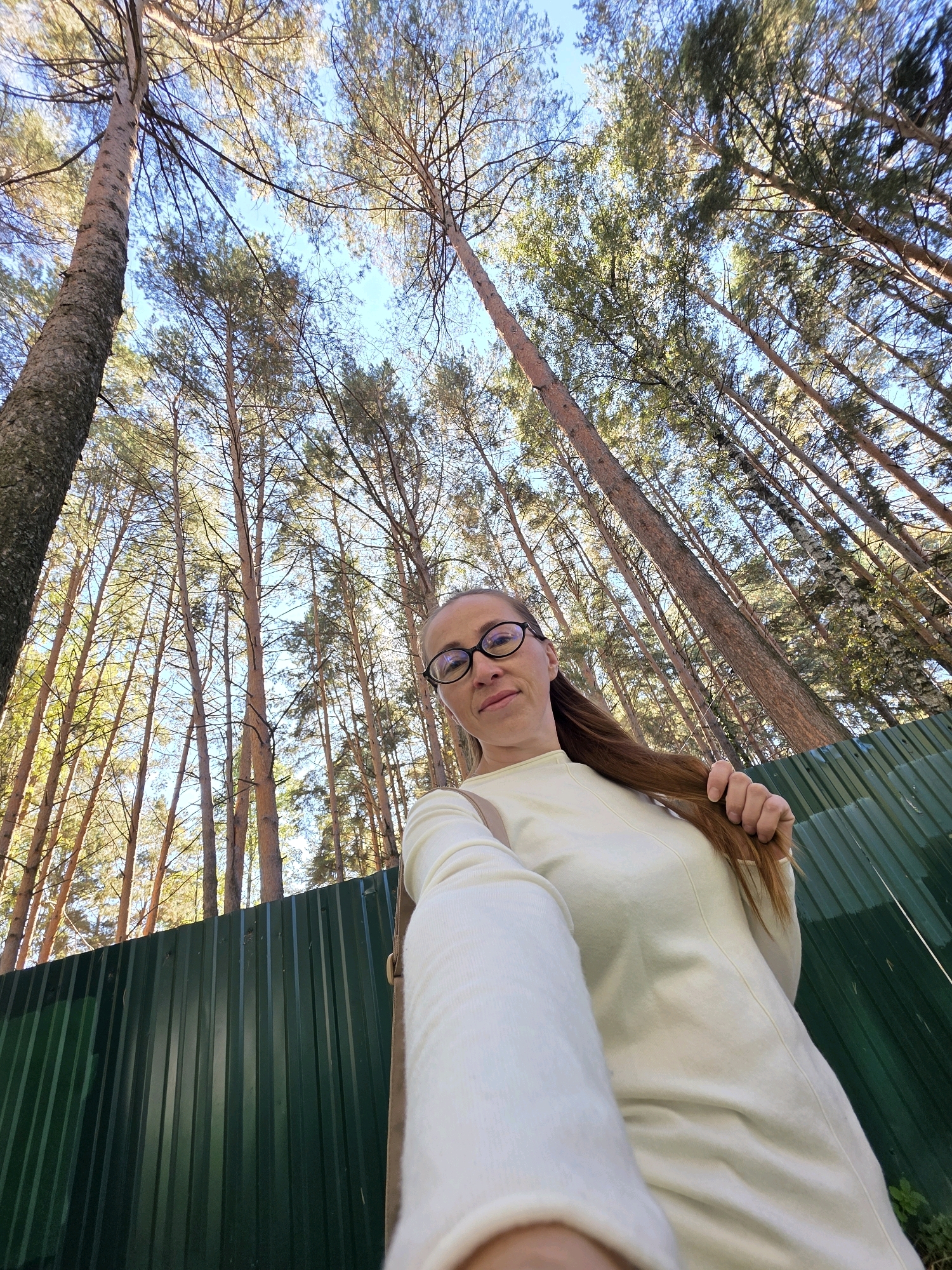 A woman with glasses and long brown hair is taking a selfie in a forest. She is wearing a light-colored sweater. Behind her, tall pine trees reach towards a clear blue sky. A green metal fence is visible in the foreground. The scene appears to be a park or natural area. The image was taken in Chaikovsky, Russia.