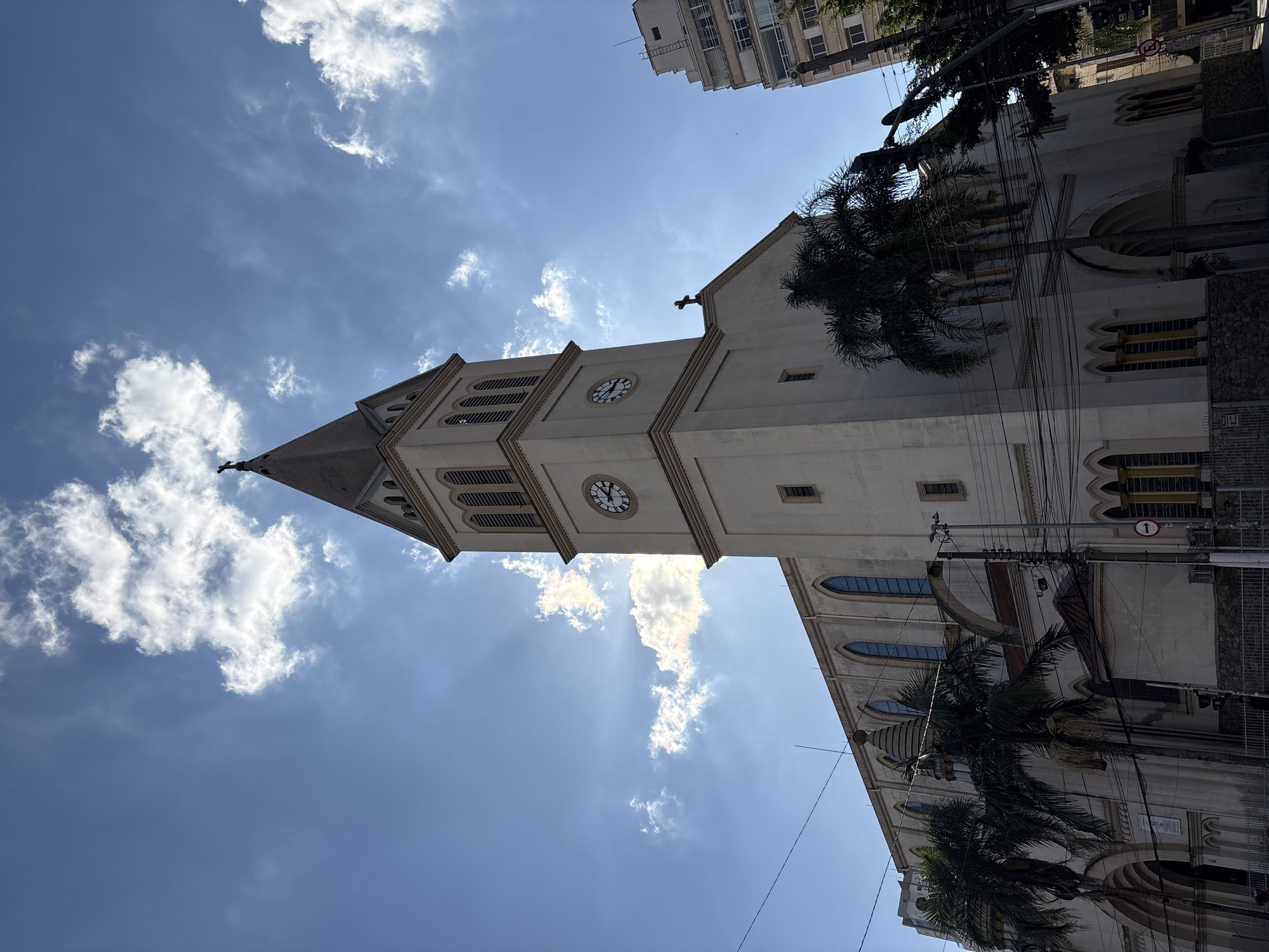 A low-angle, wide shot captures the facade of a large, light-colored church with a prominent bell tower and a tall, pointed steeple topped with a cross. The church features arched windows and intricate architectural details. In the foreground, palm trees partially obscure the lower sections of the building. Above, a bright blue sky is dotted with white, fluffy clouds, and sunbeams emanate from behind one of the clouds, creating a dramatic effect. A modern, multi-story building with visible windows stands to the right of the church, contrasting with the older architecture. Electrical wires crisscross the scene, and a traffic sign with a red circle and a white line is visible on the right. The image appears to be taken on a sunny day in the city of São Paulo, Brazil, possibly on Paulista Avenue due to the presence of both older religious structures and modern high-rise buildings. There are no discernible people in the frame.