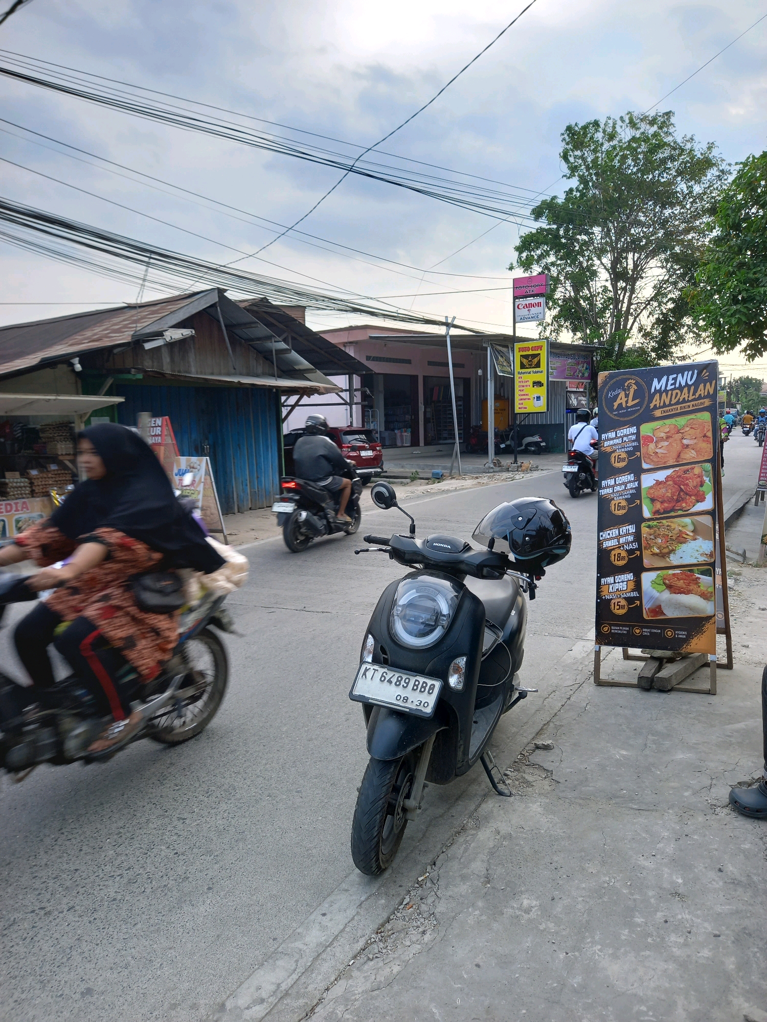 A street scene in Samarinda, Indonesia, is captured under an overcast sky. In the foreground, a black scooter is parked on the side of the road, its license plate reading "KT 6489 BBB 09-30". A menu board for "Kedai AL ANDALAN" stands next to it, displaying pictures of food items and prices. The menu lists "AYAM GORENG", "AYAM GORENG TERI", "CHICKEN KATSU SAMBAL MATAH", and "AYAM GORENG KIPAS".
To the left, a woman wearing a black hijab and a patterned orange and brown top rides a motorcycle, with a passenger behind her also wearing a black hijab. They are in motion, blurred due to speed. The road is paved, with a concrete curb and some debris.
In the background, buildings line the street, including what appears to be a shop with a blue facade and a brown corrugated metal roof. There are also advertisements and signs visible, including one for "Canon" and another for "FOTO COPY". Several other motorcycles and a red car are also present on the road, with people riding them. Trees and utility wires are also part of the urban landscape. The overall impression is a typical street view in a bustling Indonesian city.