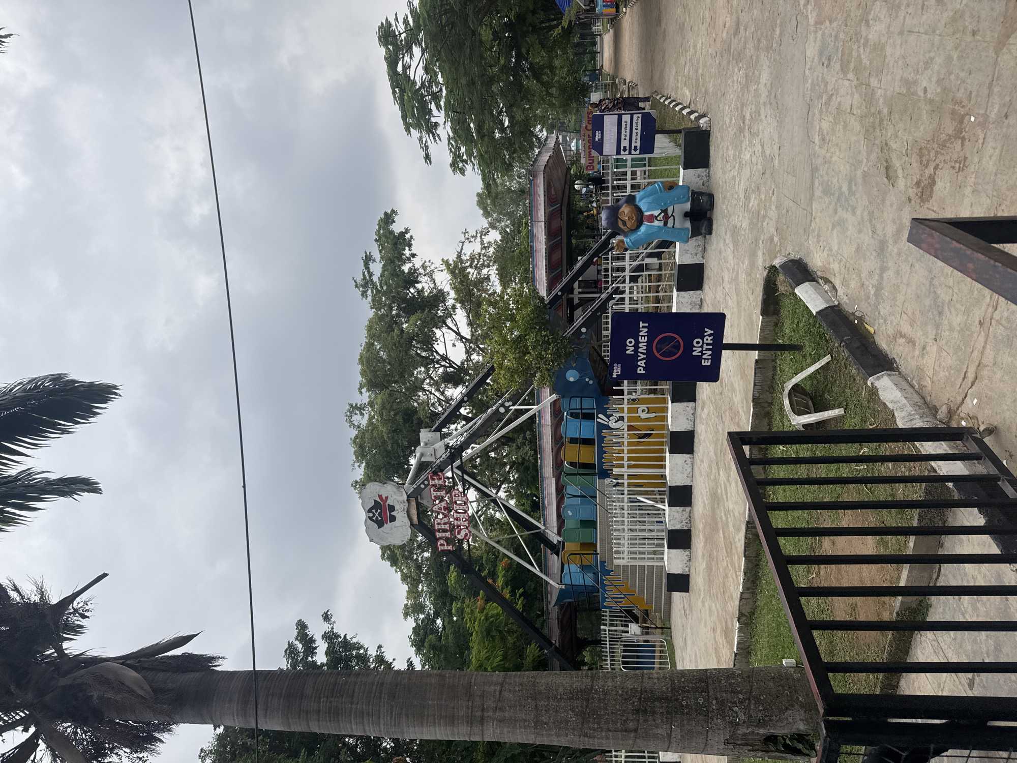 The image shows a pirate ship-themed amusement park ride at a location in Abuja, Nigeria. The ride is unoccupied and features a sign stating "NO PAYMENT, NO ENTRY." There is a painted pirate figure standing near the entrance to the ride. A black metal gate is visible in the foreground, and there is a palm tree on the left side of the frame. In the background, there are trees and other structures within the park, including signage directing visitors to "Paintball" and "Horse Riding". The sky is overcast.